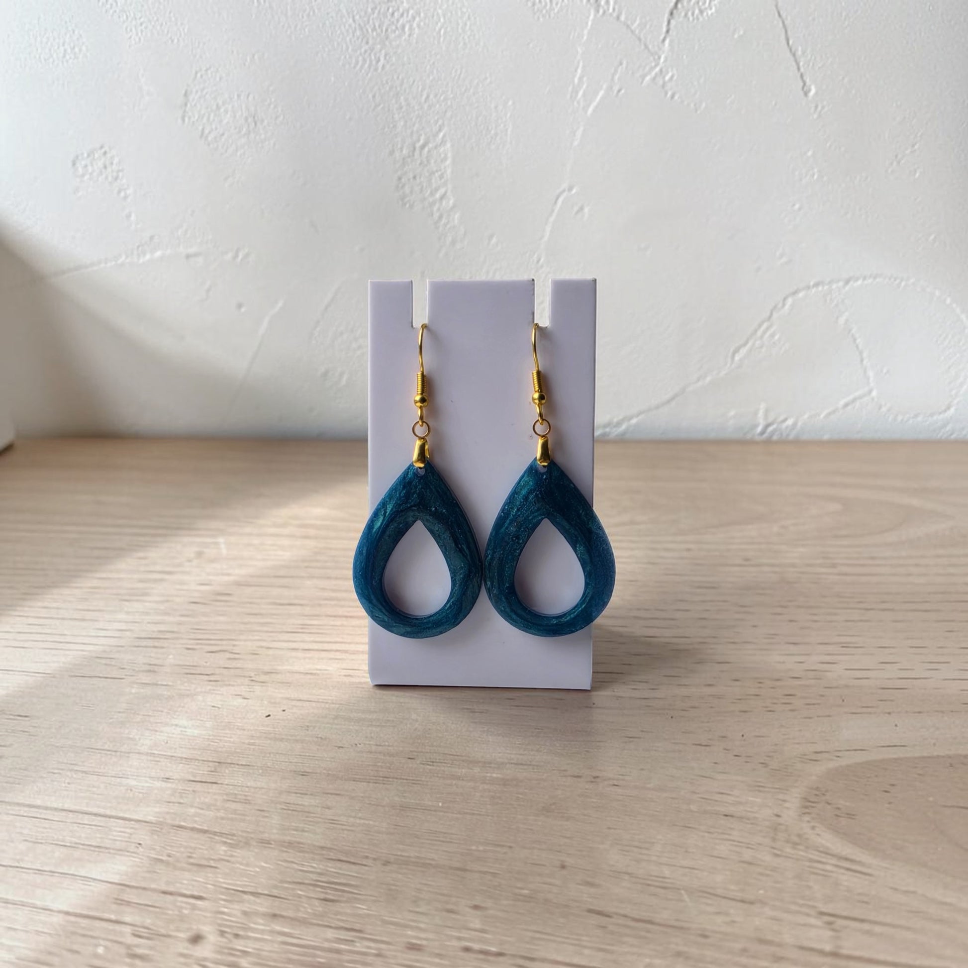 Mirabilia Boutique’s Mermaid Dust Teardrop Earrings with gold hooks are shown on a white stand against a textured wall—an ideal choice for mermaid jewelry lovers.