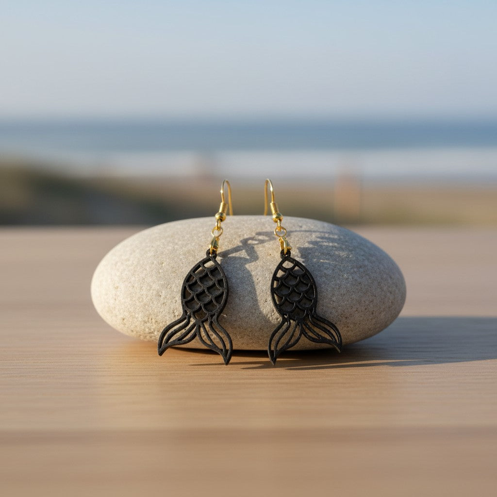A pair of Mermaid Tail Earrings | Lasercut Walnut Wood by Mirabilia Boutique hang from a smooth stone on wood, with the beach and ocean blurred in the background—ideal coastal jewelry for ocean lovers.