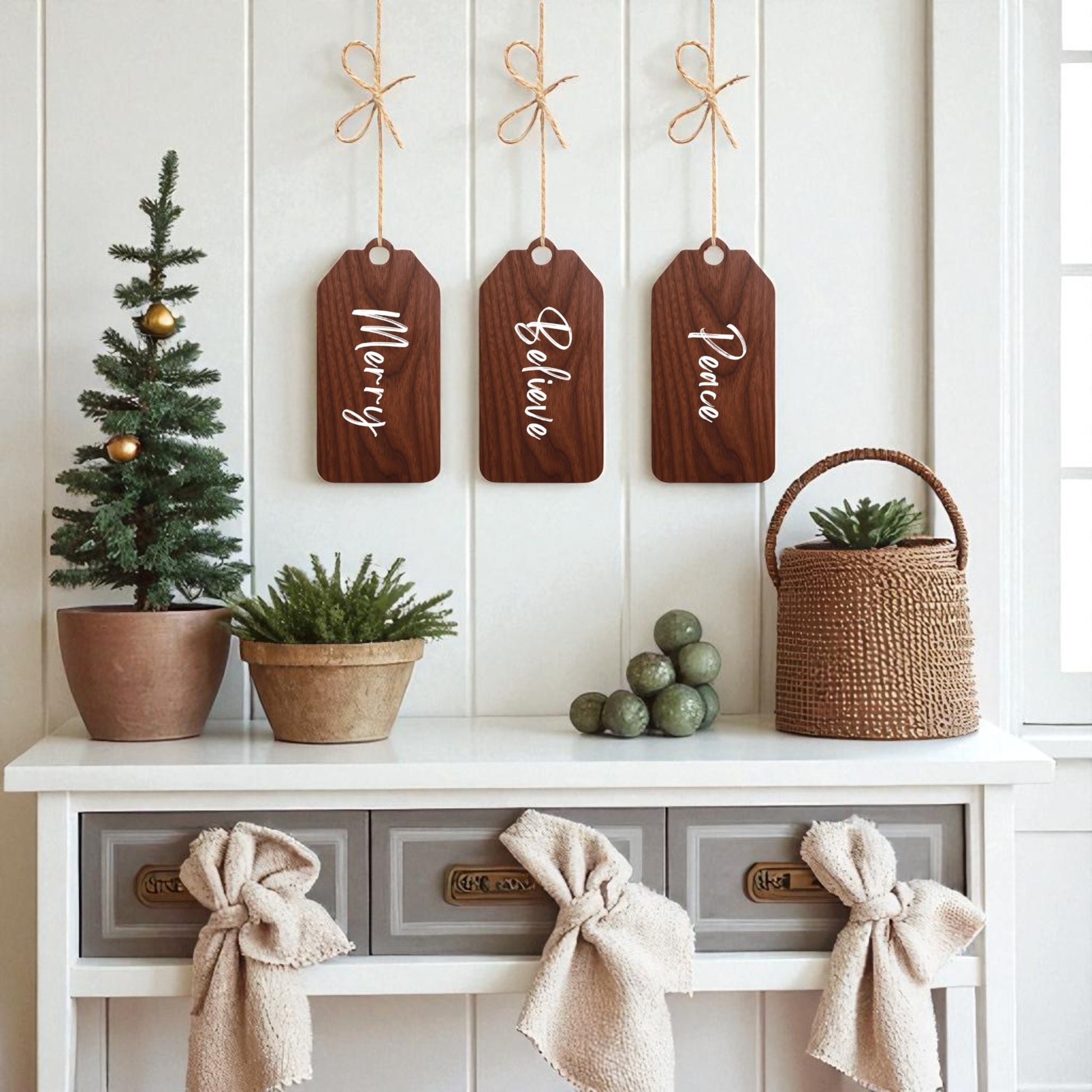 A white table with drawers displays rustic holiday decor, featuring a small Christmas tree and “Merry Believe Peace” wooden tags by Mirabilia Boutique hanging above. Potted plants, bows, and a woven basket add festive charm.