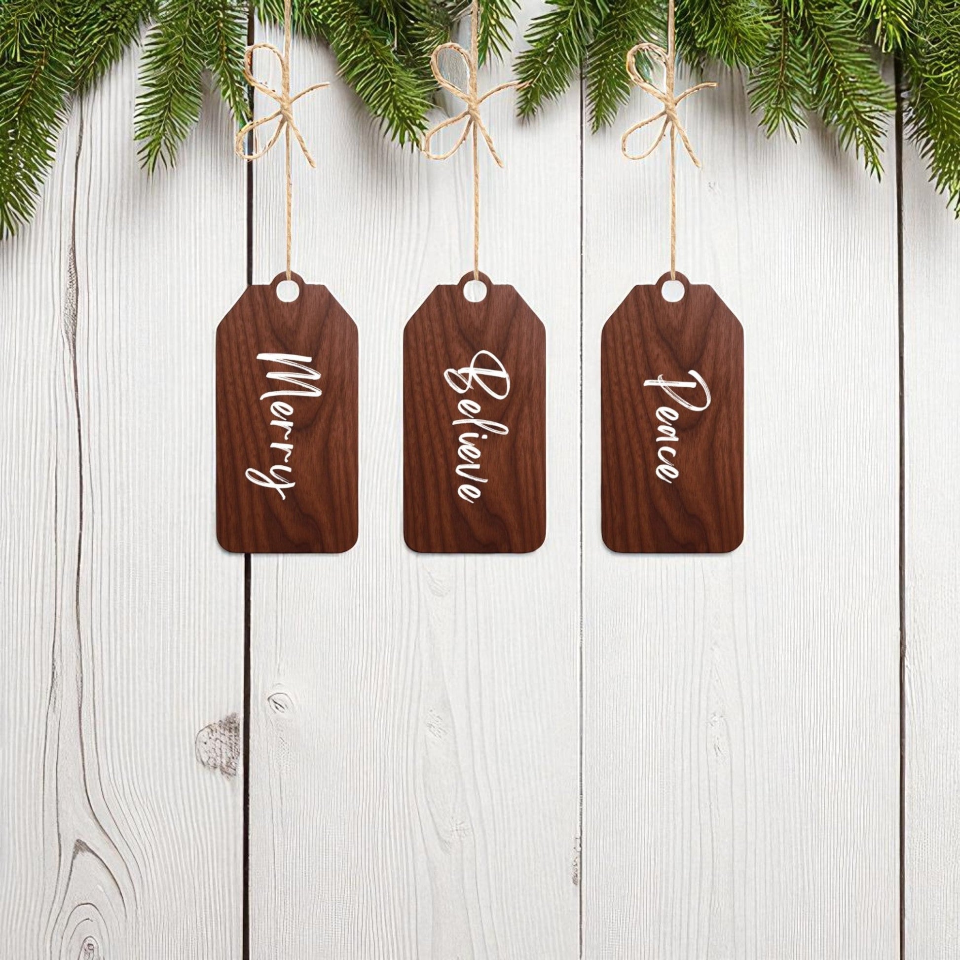 Mirabilia Boutique’s Merry Believe Peace Tags set features three 5x2 rustic wood ornaments with festive sayings, hanging from strings under evergreen branches for charming Christmas décor.