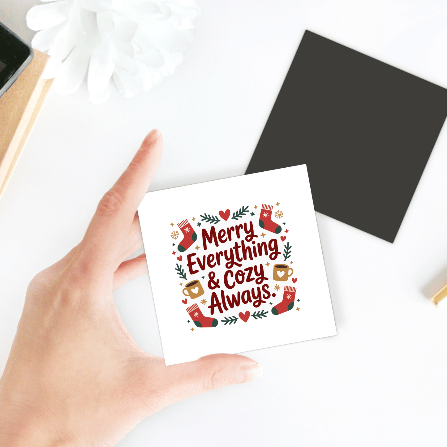 A hand holds the Mirabilia Boutique "Merry Everything & Cozy Always" Holiday Fridge Magnet, featuring festive illustrations—perfect as a stocking stuffer or cozy holiday decor. A black square and white flower are in the background.