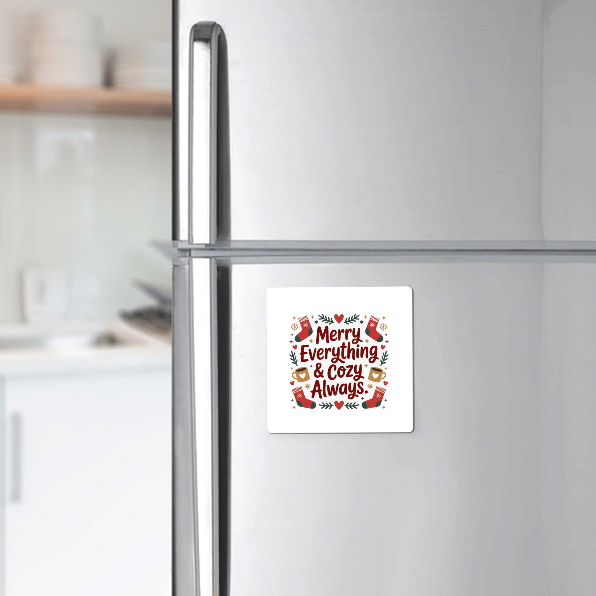 The Merry Everything & Cozy Always Magnet by Mirabilia Boutique decorates a stainless steel fridge with festive stockings, mugs, and greenery. A kitchen counter with dishes appears in the background.