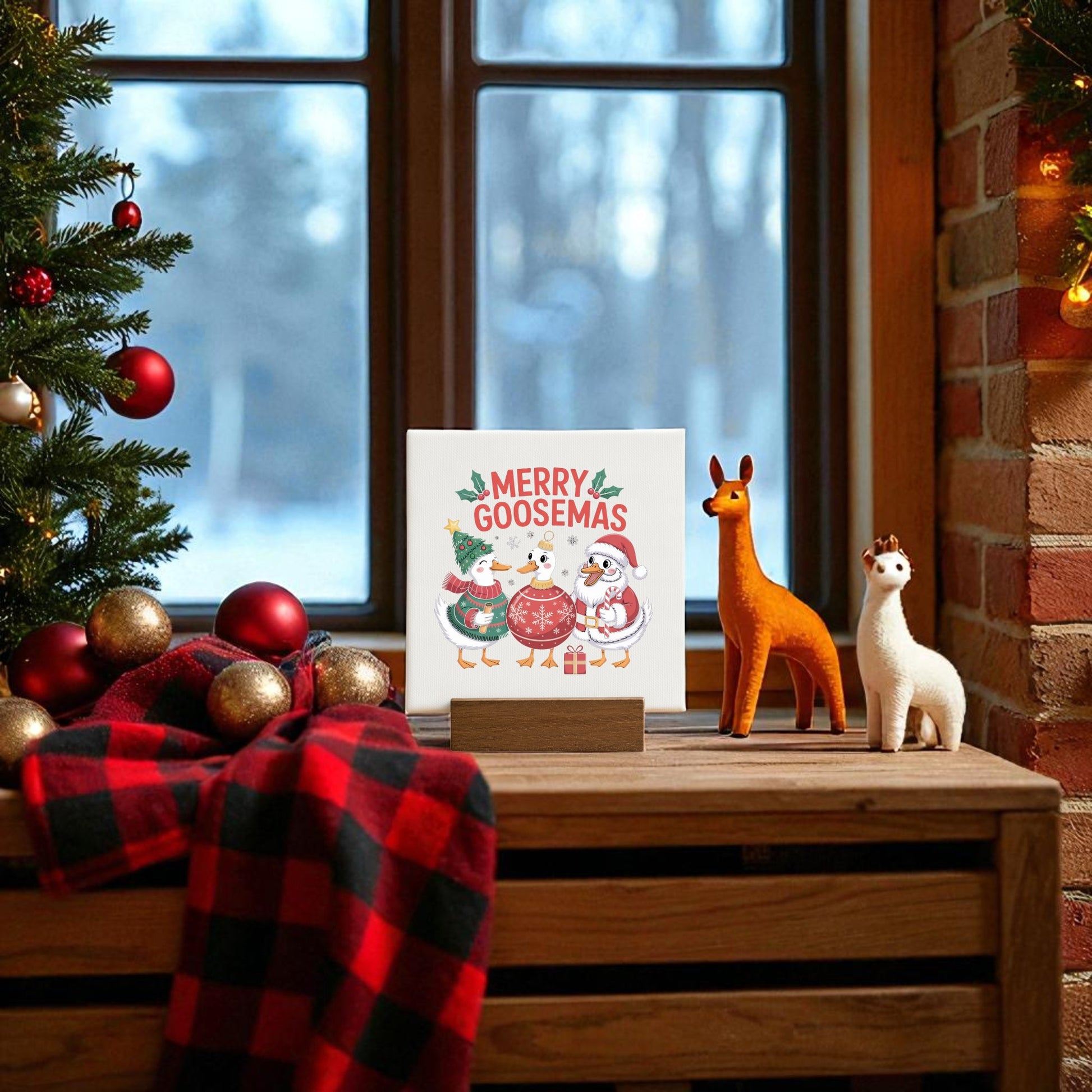 The Merry Goosemas Mini Sign by Mirabilia Boutique sits on a wooden table near a window, flanked by llama figurines, a plaid blanket, ornaments, and a festive tree—a quirky holiday gift and charming decor with snowy views outside.