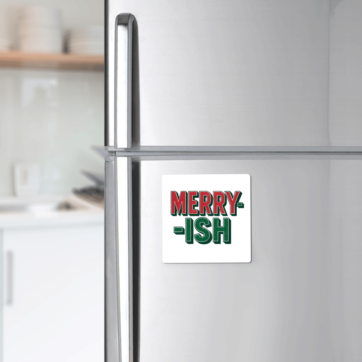 A white refrigerator features the Merry-ish Magnet by Mirabilia Boutique, a funny holiday fridge decor with bold red and green lettering; a blurred kitchen appears in the background.