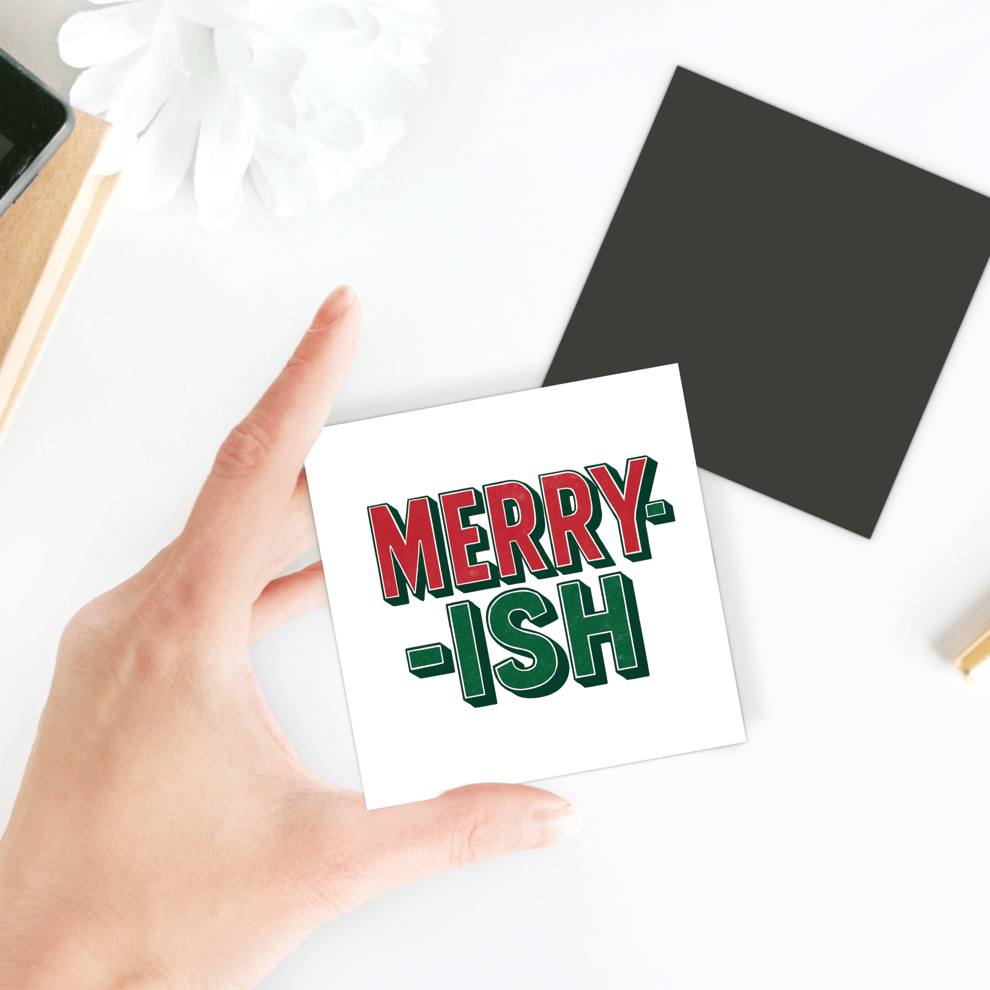 A hand holds a "MERRY-ISH" card next to the Merry-ish Magnet by Mirabilia Boutique—a black, square, funny holiday fridge decor—on a white surface with a white flower and desk items nearby.