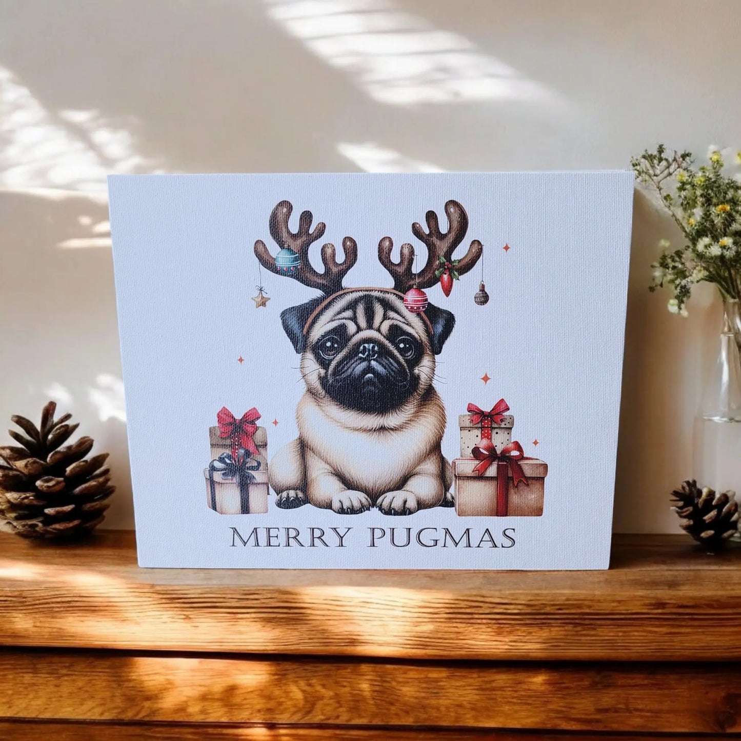 A framed holiday decor piece from Mirabilia Boutique sits on a wooden shelf. The Merry Pugmas 10x8 Canvas Sign features a cute pug wearing reindeer antlers surrounded by wrapped presents, with the cheerful text "MERRY PUGMAS" displayed beneath. Pinecones and a small plant are placed beside the festive print.