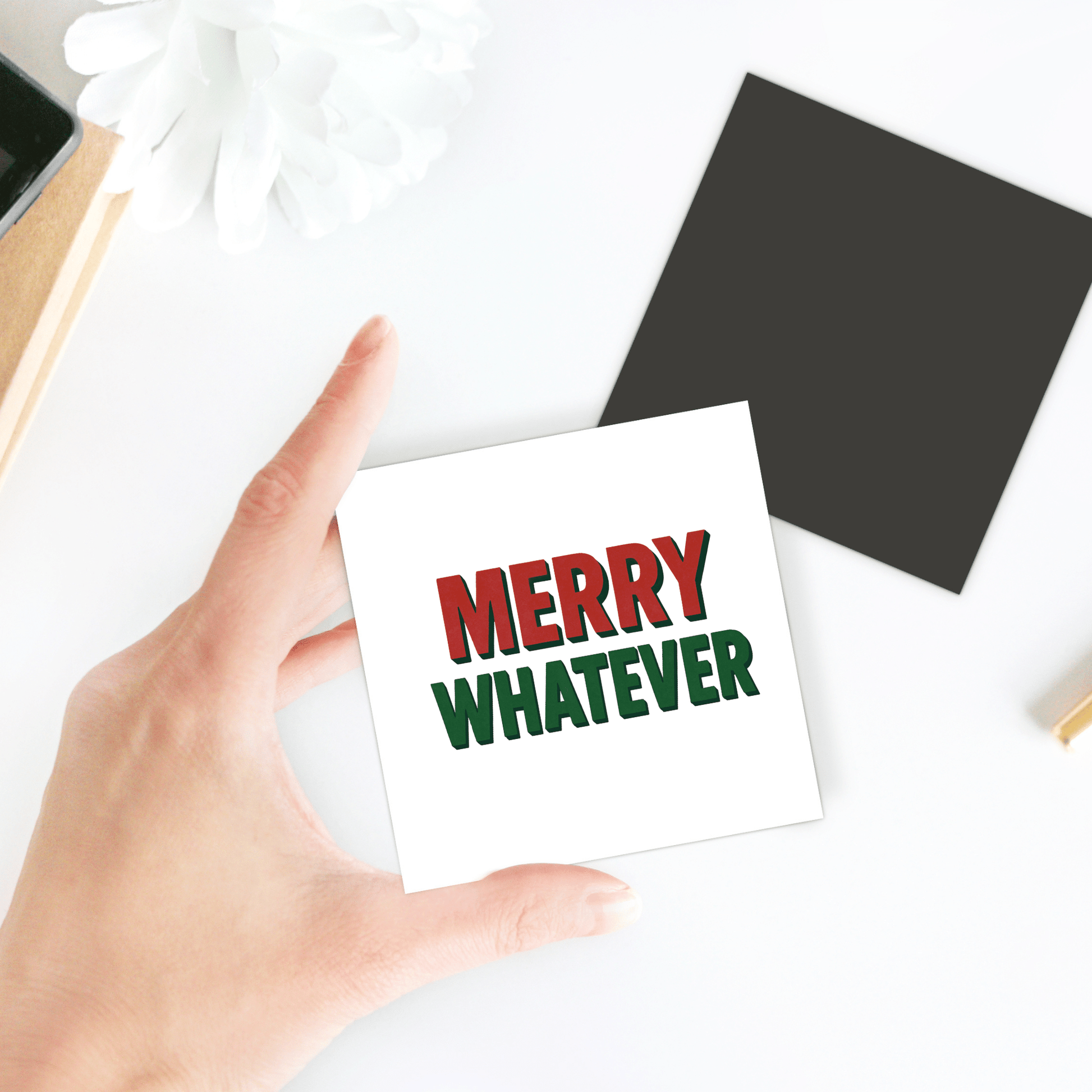 A hand holds the Merry Whatever Magnet by Mirabilia Boutique—a funny minimalist Christmas gift. In the background, there's a black square card, a white flower, and a notebook on the white table.