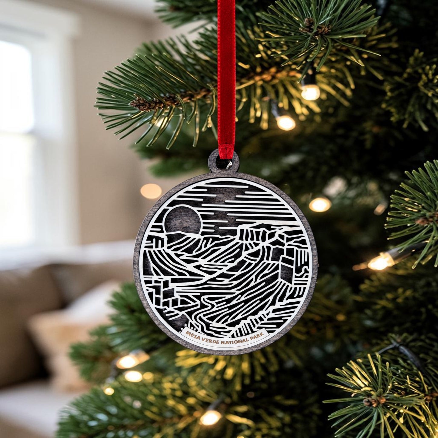 A Mesa Verde National Park Line Art Christmas Ornament by Mirabilia Boutique, showcasing intricate black-and-white cliff dwellings, hangs from a Christmas tree branch adorned with lights, suspended by a red ribbon.