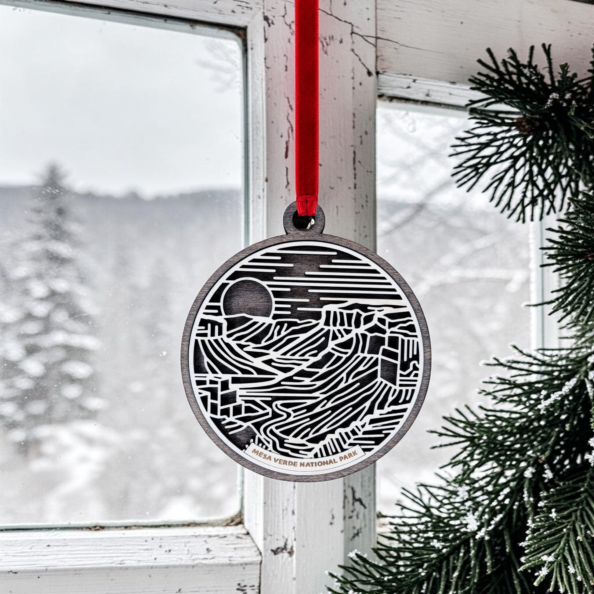 The Mesa Verde National Park Line Art Christmas Ornament by Mirabilia Boutique, featuring a red ribbon, hangs near a snowy window framed by green pine branches—a graceful tribute to the park’s iconic cliff dwellings with a winter landscape visible outside.