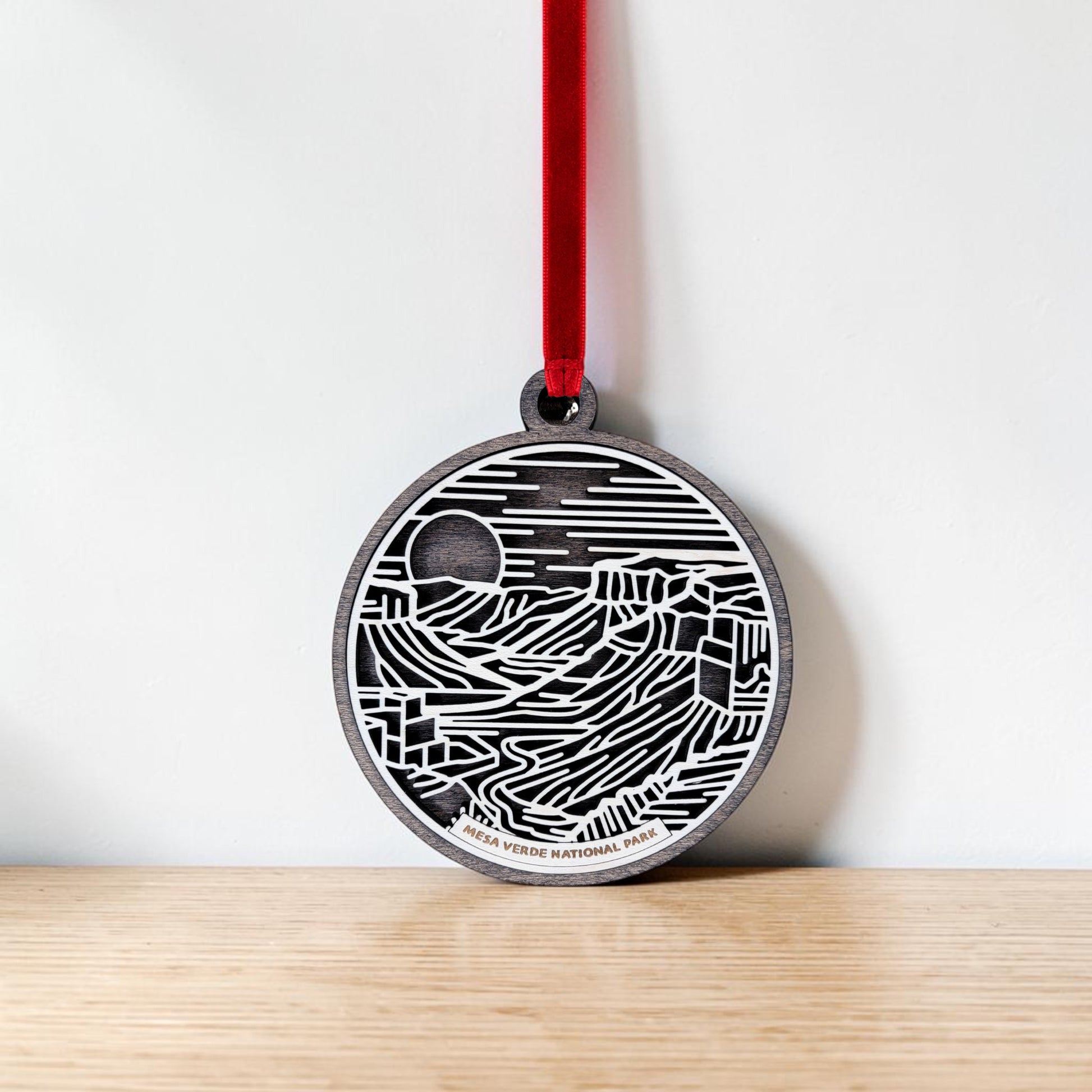 The Mesa Verde National Park Line Art Christmas Ornament by Mirabilia Boutique features a round, black and white design with abstract landscape lines, a sun motif, and subtle nods to the ancient cliff dwellings. Hanging from a red ribbon above a wooden surface against a plain white wall, this handcrafted wooden keepsake is ideal for anyone who treasures the history and beauty of Mesa Verde.