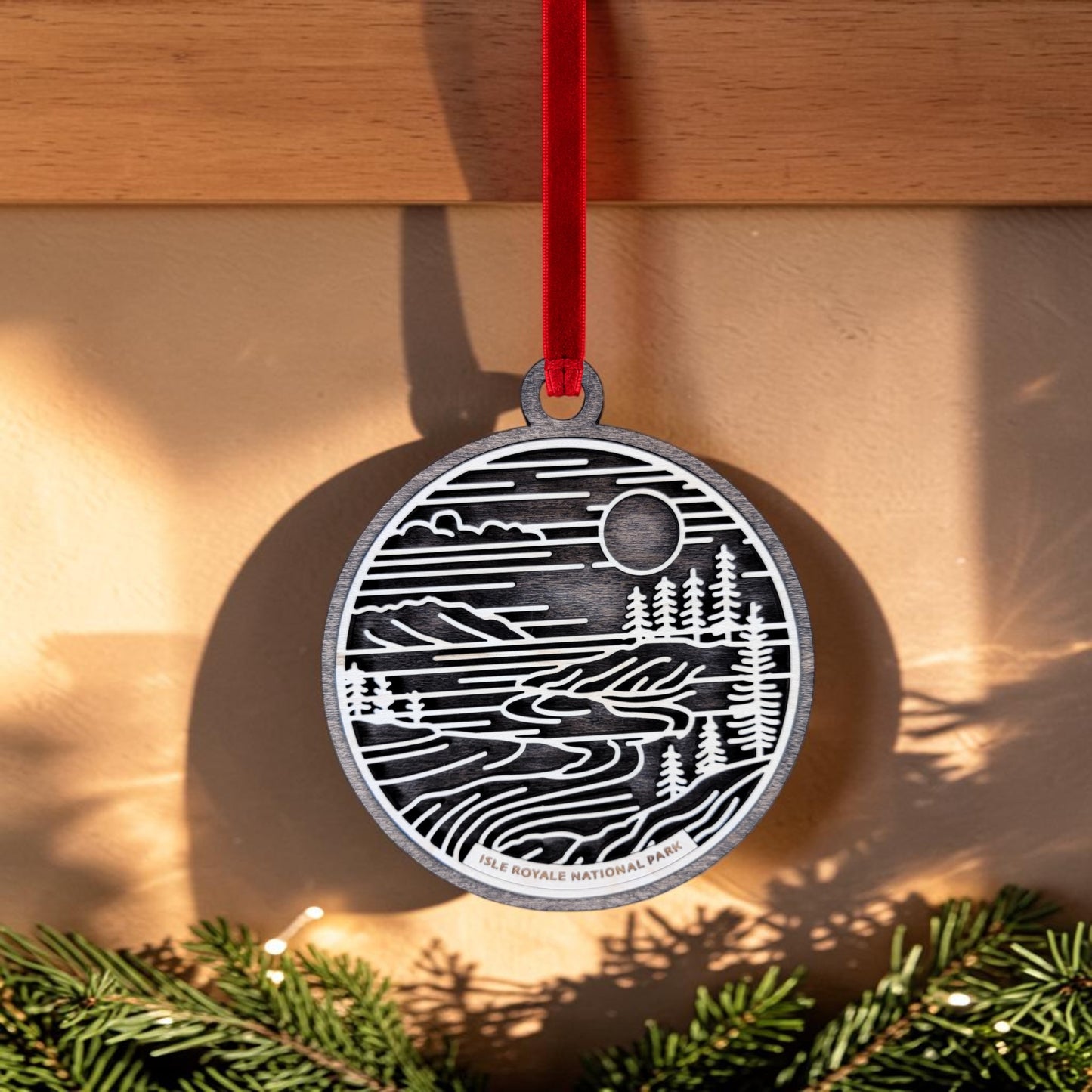 The Isle Royale National Park Line Art Christmas Ornament by Mirabilia Boutique features a black and white landscape design on an oval wooden ornament. It hangs from a red ribbon, with sunlight casting subtle shadows and pine branches accenting the bottom edge. The ornament is inscribed with "ISLE ROYALE NATIONAL PARK," beautifully capturing the tranquil spirit of Lake Superior's remote wilderness.