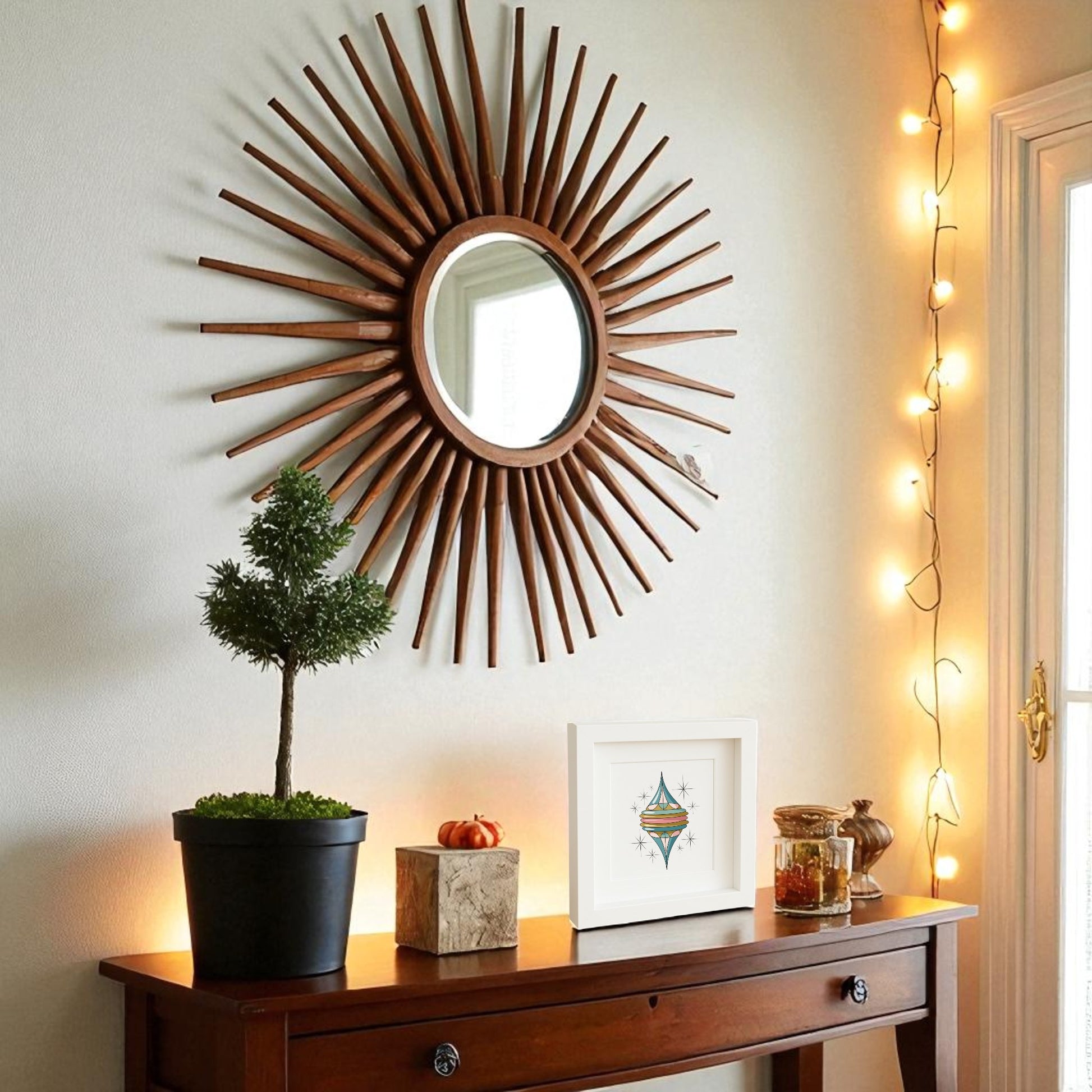 A wooden sunburst mirror hangs above a table with a potted plant, mini pumpkin, and the Retro Icicle Ornament Art by Mirabilia Boutique. String lights drape the wall and window for a festive, mid-century holiday vibe.