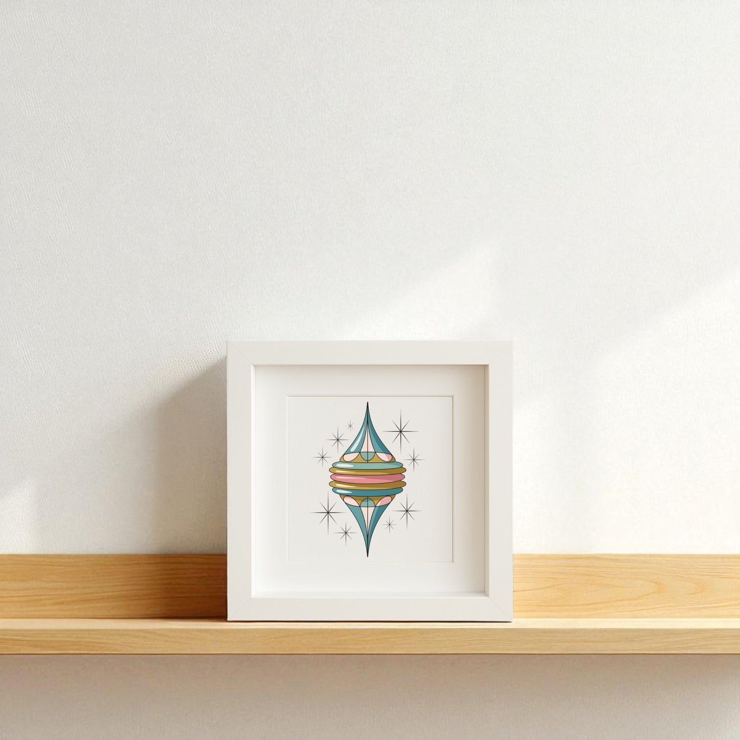 The Retro Icicle Ornament Art by Mirabilia Boutique, a mid-century Christmas shadowbox with colorful geometric design, rests on a light wooden shelf, adding retro holiday charm to a plain white wall.