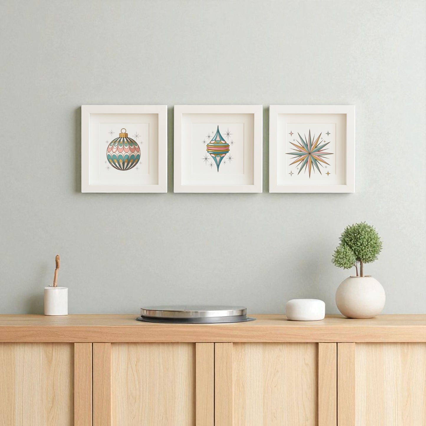 A minimalist wooden sideboard displays a round tray, white pot, and plant. Above, three Mirabilia Boutique "Mid-Century Round Ornament Art | Retro Holiday Shadowbox" prints add retro holiday charm to the light gray wall.