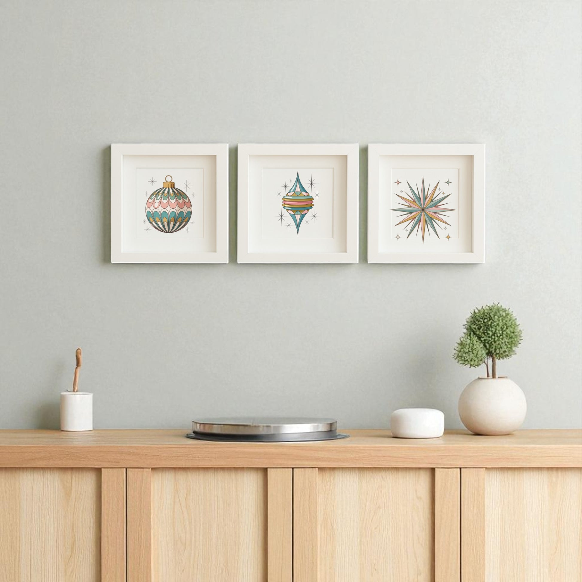 The Retro Ornament Trio Mid-Century Holiday Wall Art Set by Mirabilia Boutique features three framed prints with teal, orange, and gold retro shapes above a light wood cabinet adorned with vintage Christmas touches on a light gray wall.