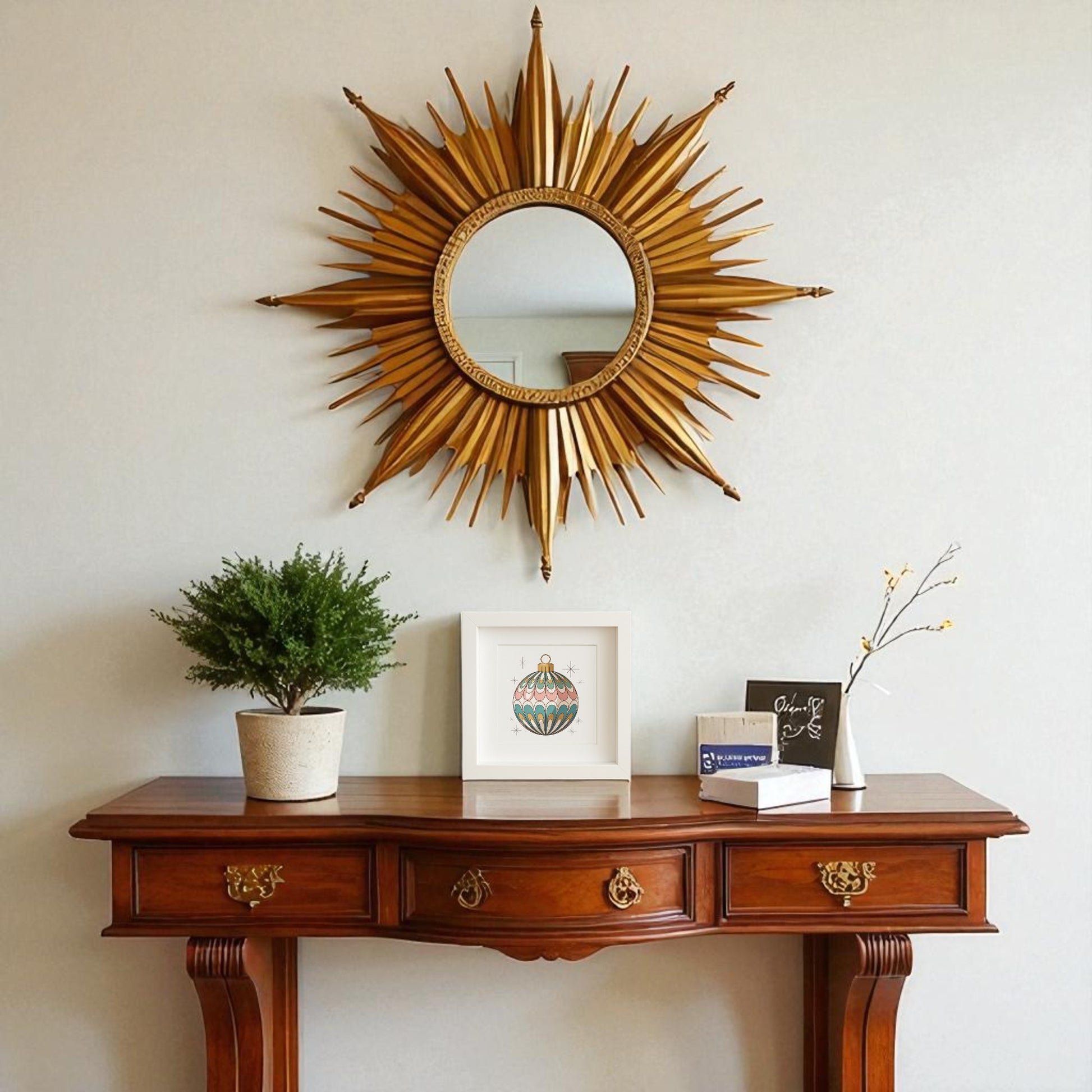 A wooden console table displays a potted plant, books, and a vase while vintage art from Mirabilia Boutique’s "Mid-Century Round Ornament Art | Retro Holiday Shadowbox" sits beside them. Above hangs a round gold sunburst mirror on the plain light wall.