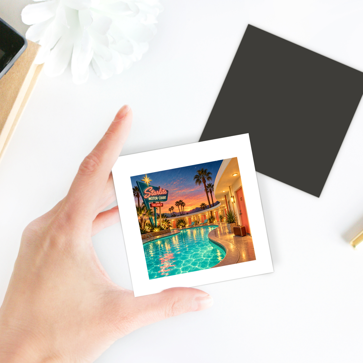 A hand holds the Arrival at the Starlite Magnet | Chrome & Starlight by Mirabilia Boutique, featuring a sunset motel scene—ideal roadside decor for your fridge. Desk items and a blank black card sit in the background.
