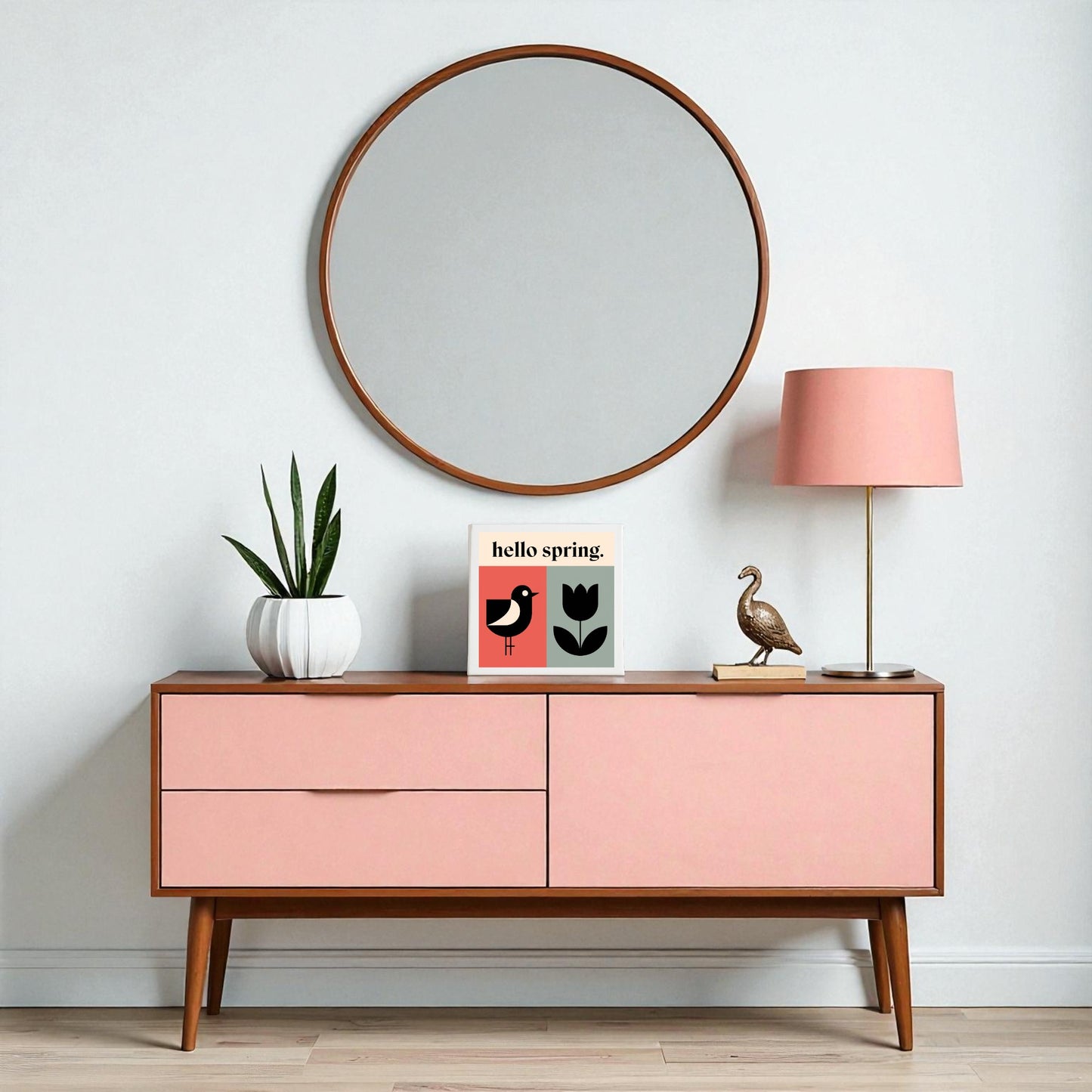 A modern sideboard with pink drawers displays a white potted plant, the Mirabilia Boutique "Hello Spring Canvas – Bird & Tulip Wall Art," cheerful home décor, a brass bird statue, and a pink lamp. Above, a large round mirror adds stylish wall decor.