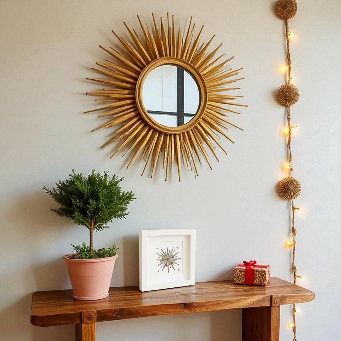 A wooden table with a small potted plant, a framed picture, and a wrapped gift sits against a wall. Above, the Mirabilia Boutique Atomic Starburst Ornament Art | Retro Holiday Shadowbox adds stylish atomic flair.