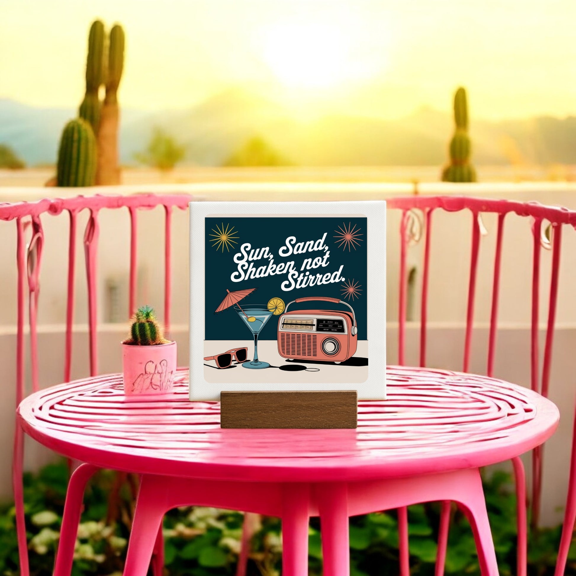 A pink mid-century modern table displays the "MCM Sun, Sand, Shaken Not Stirred Mini Canvas – 4x4 Retro Sign" by Mirabilia Boutique, featuring a chic retro design with a car and cactus.