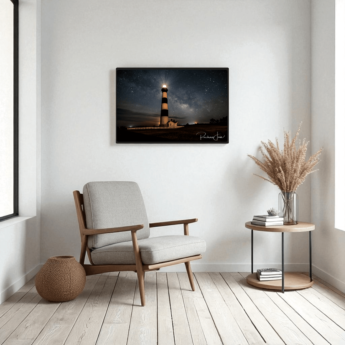 A modern living room with a light armchair, round side table with dried grasses, and Mirabilia Boutique’s “Milky Way at Bodie Lighthouse” framed art print showcasing an Outer Banks nightscape. Natural light streams in from a left window.
