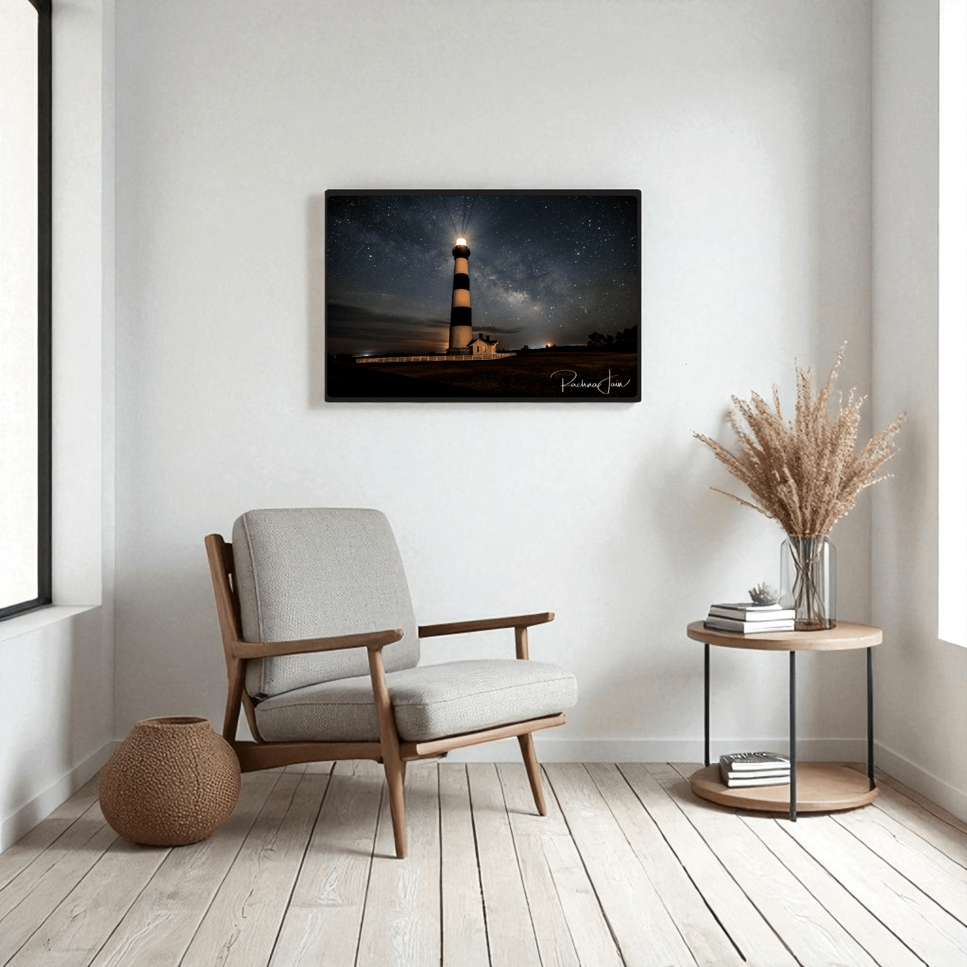 A modern living room with a light armchair, round side table with dried grasses, and Mirabilia Boutique’s “Milky Way at Bodie Lighthouse” framed art print showcasing an Outer Banks nightscape. Natural light streams in from a left window.