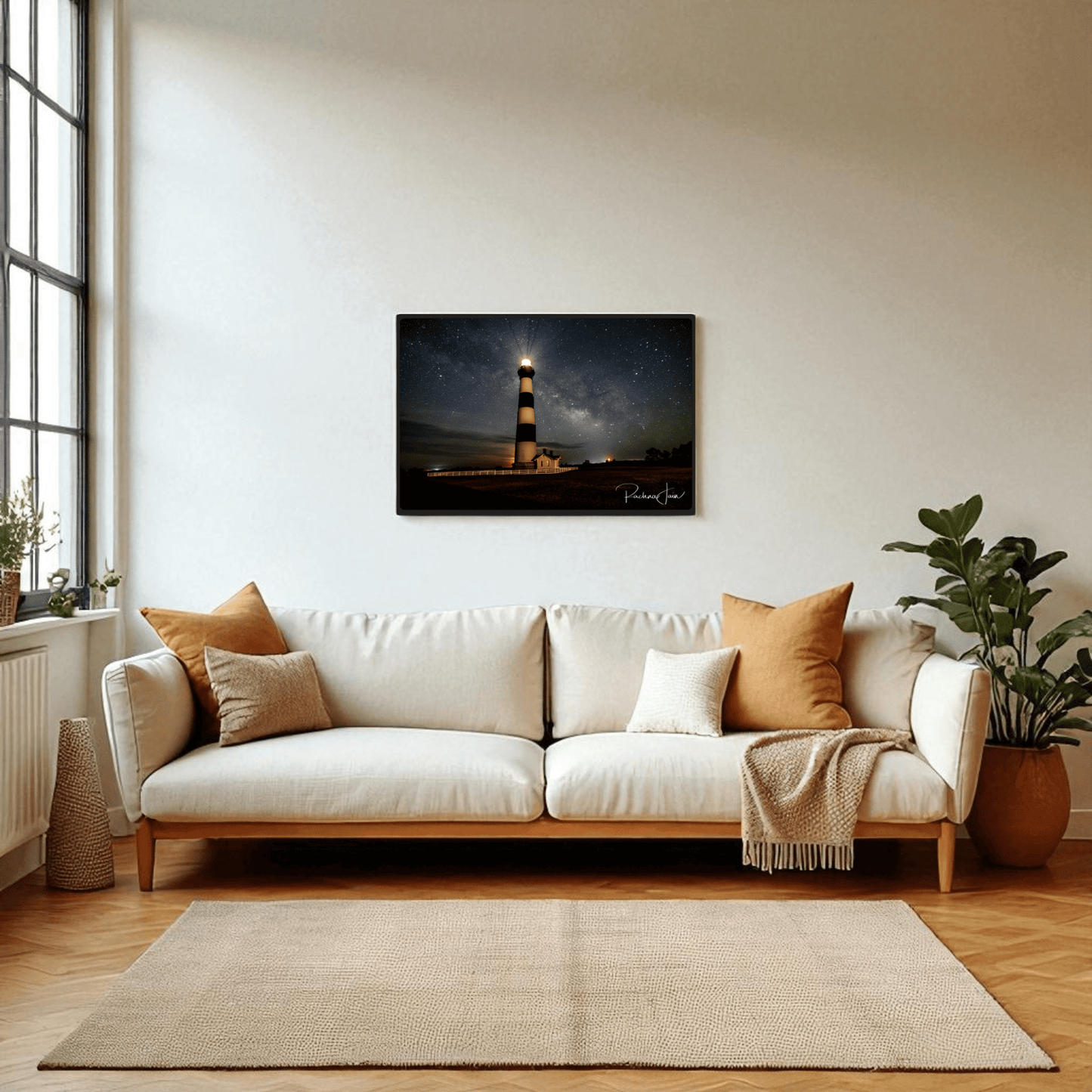 A bright, cozy living room features a white sofa with brown and beige cushions, a blanket, green potted plants, and Mirabilia Boutique’s "Milky Way at Bodie Lighthouse" Outer Banks nightscape framed art print illuminated by natural light.