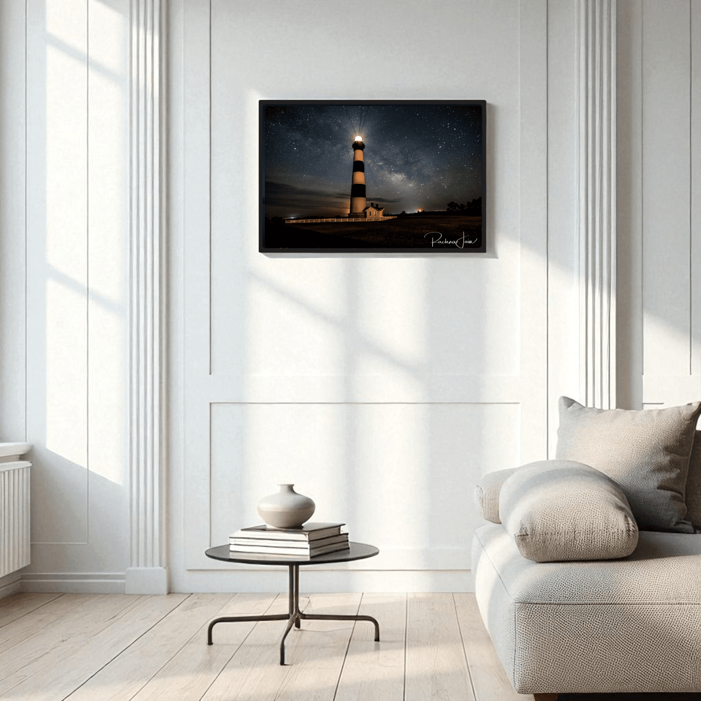 A minimalist living room features a light sofa, round coffee table, and vase. On the white-paneled wall hangs Mirabilia Boutique’s “Milky Way at Bodie Lighthouse” framed art print, capturing an Outer Banks nightscape.
