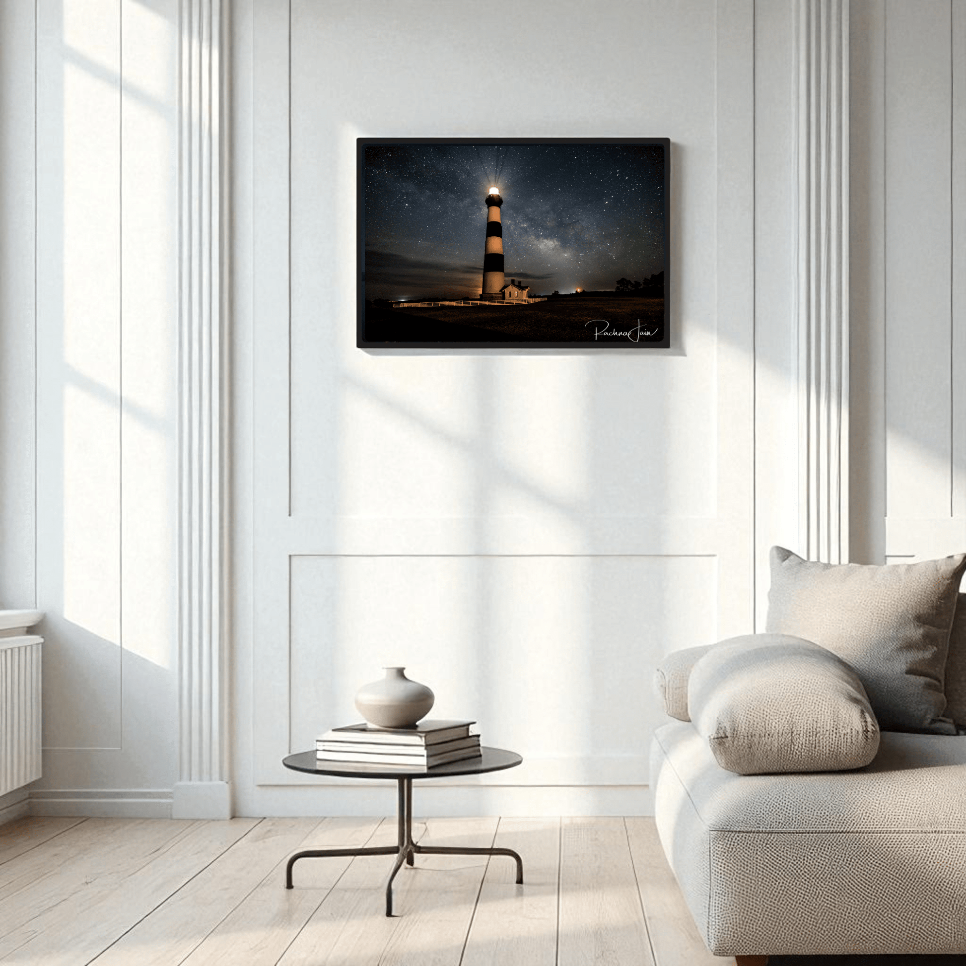 A minimalist living room features a light sofa, round coffee table, and vase. On the white-paneled wall hangs Mirabilia Boutique’s “Milky Way at Bodie Lighthouse” framed art print, capturing an Outer Banks nightscape.