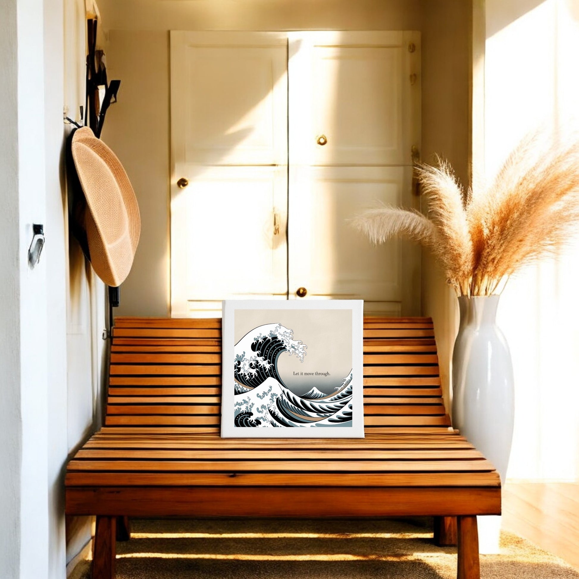 The Let It Move Through Canvas – Bold Ocean Calm Art by Mirabilia Boutique rests on a wooden bench. A large vase with pampas grass and a wall hook with a straw hat complete the look, as sunlight fills this resilient, cozy hallway.