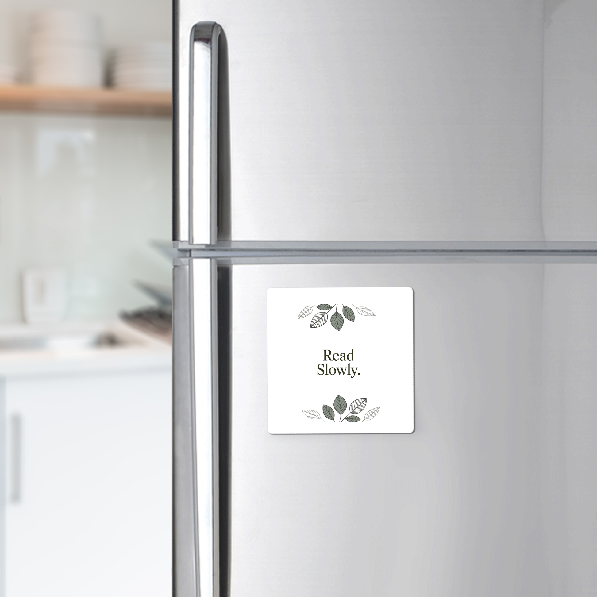 The Read Slowly Book Lover Magnet by Mirabilia Boutique—a white square magnet with green leaves and the words "Read Slowly."—adds literary charm to any silver refrigerator. White dishes are softly blurred in the background.