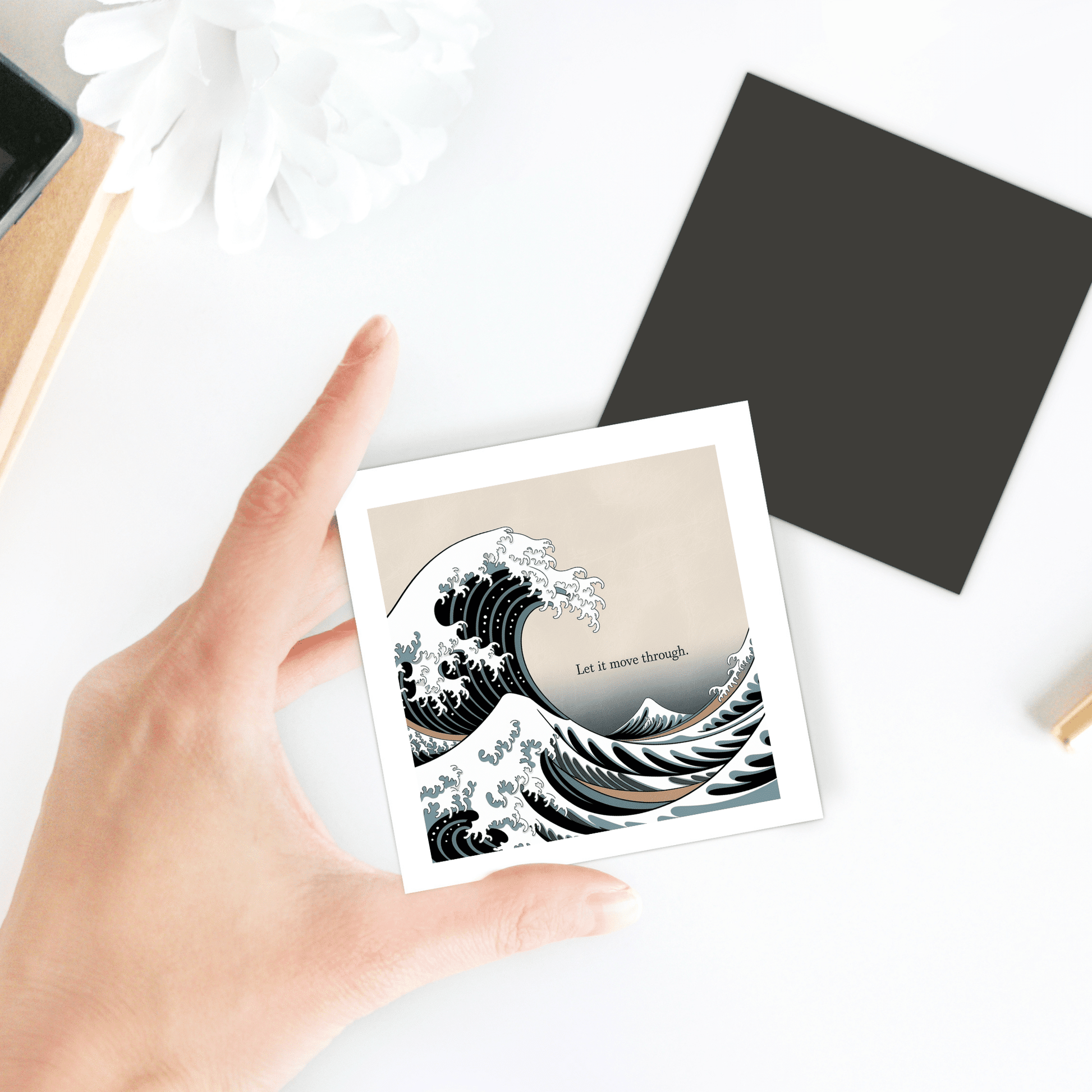 A hand holds the "Let It Move Through Magnet – Storm & Stillness Quote" by Mirabilia Boutique, featuring a wave illustration. The magnet rests on a white surface with a flower, a black square, and other nearby items.