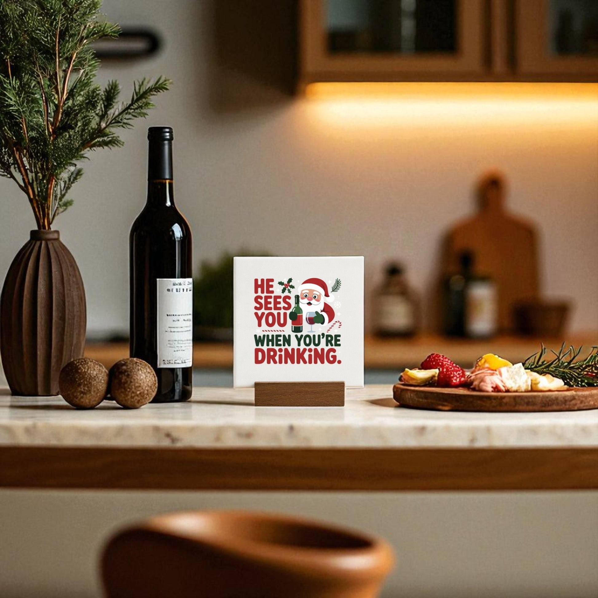 A kitchen counter features a wine bottle, a vase with greenery, fruit and cheese, and Mirabilia Boutique’s “He Sees You When You’re Drinking” funny mini Christmas sign—an ideal touch for your holiday bar cart décor.