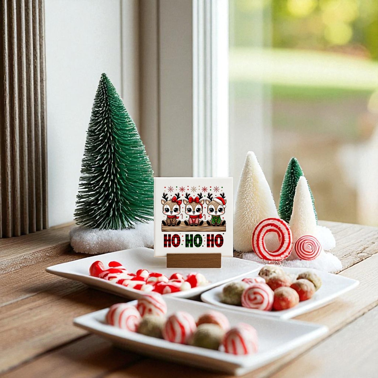 A festive holiday scene features Mirabilia Boutique’s 4x4 “Ho Ho Ho Reindeer” Mini Christmas Sign and small artificial trees, reindeer décor, and plates of red, white, and green candies arranged on a wooden surface by a window.