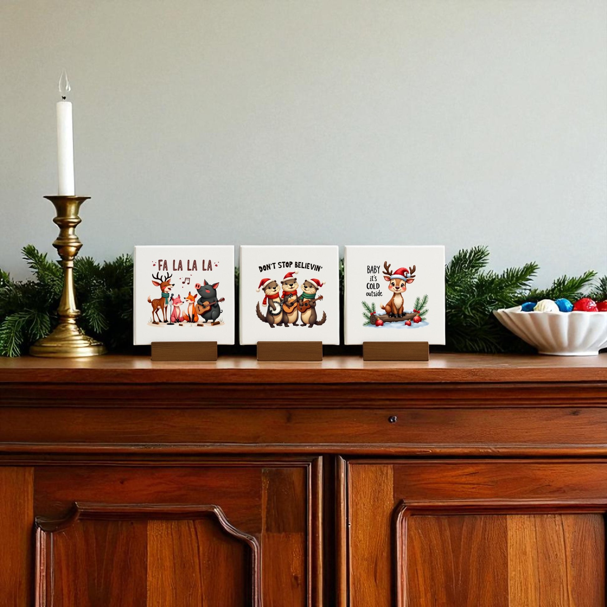 A wooden mantel adorned with greenery, a brass candlestick, ornament bowl, festive cards with reindeer and dogs in holiday wear, and Mirabilia Boutique’s Fa La La, Don’t Stop Believin, Baby It's Cold Mini Christmas Sign Trio for charming decor.
