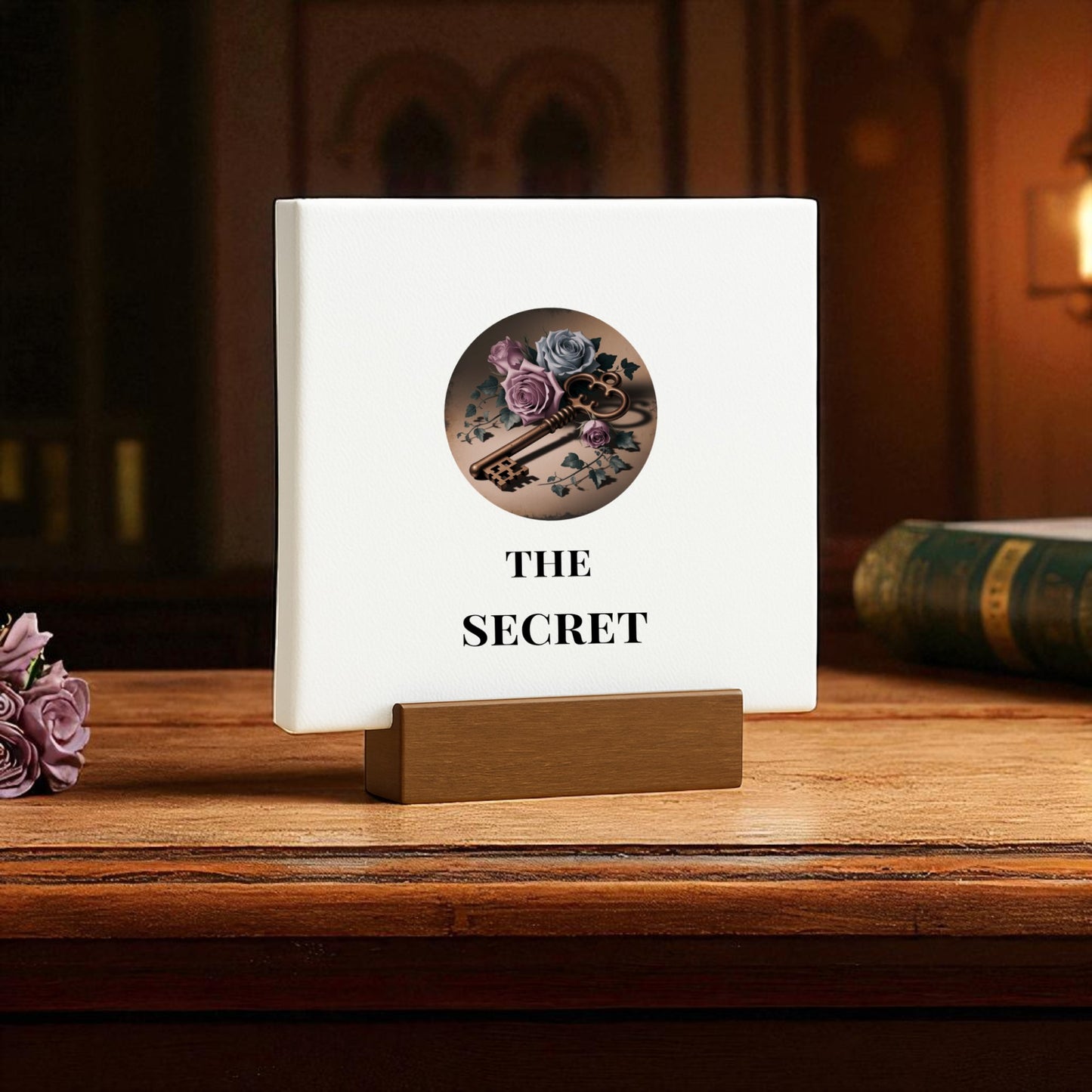 The "Mini Gothic Sign | The Secret | Dead Regency Decor" by Mirabilia Boutique stands upright on a wooden desk in a brown holder, featuring vintage skeleton key, lock, and floral art; a closed book and purple flowers sit nearby.