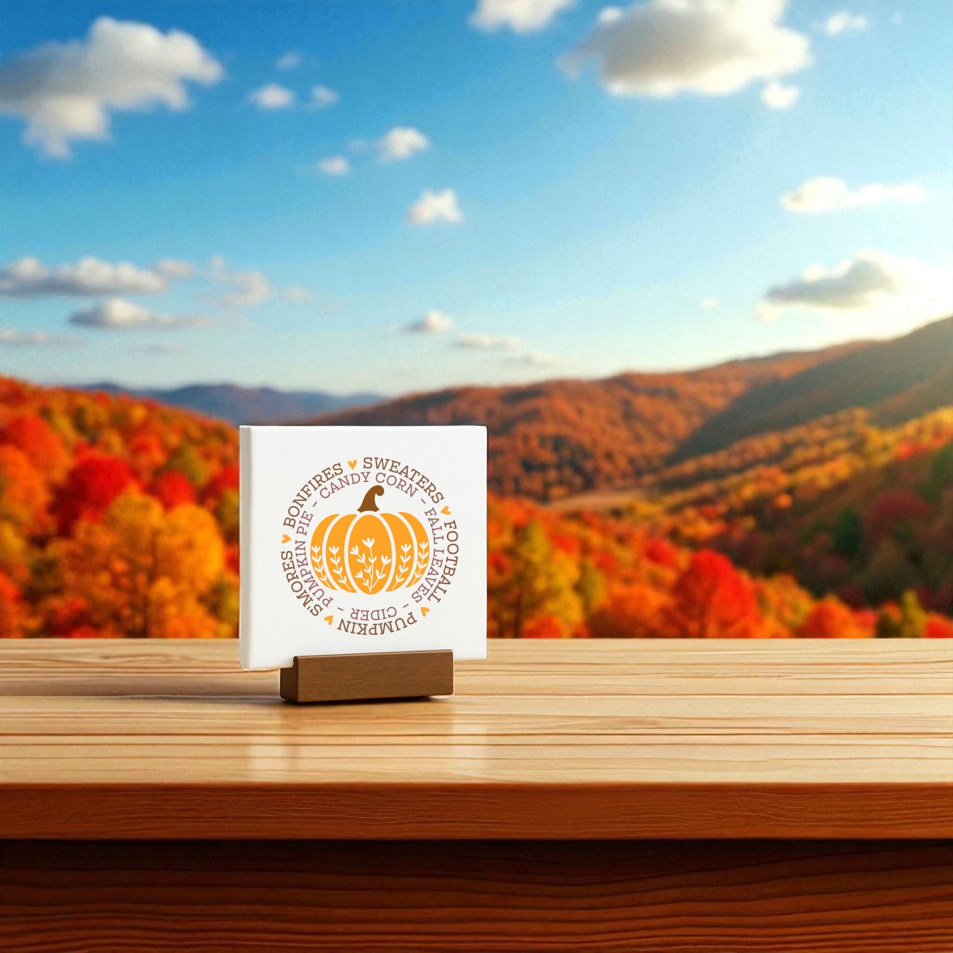The Mini Fall Sign by Mirabilia Boutique features autumn-themed words and sits on a wooden table, making cozy shelf décor. Colorful, tree-covered hills and a blue sky with clouds create the perfect seasonal backdrop.