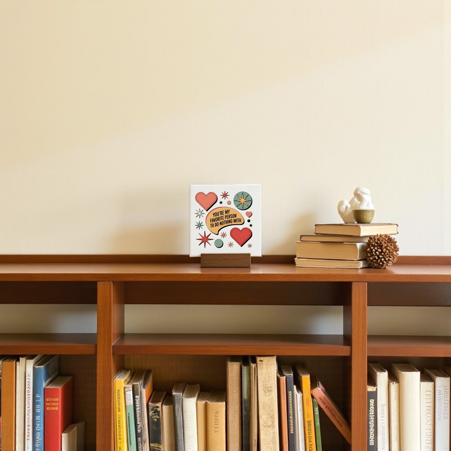The Retro Mini Love Sign by Mirabilia Boutique, featuring hearts and stars, sits on a shelf with books and a pine cone, topped by a bird figurine. This quirky decor adds charm to any cream-colored wall.