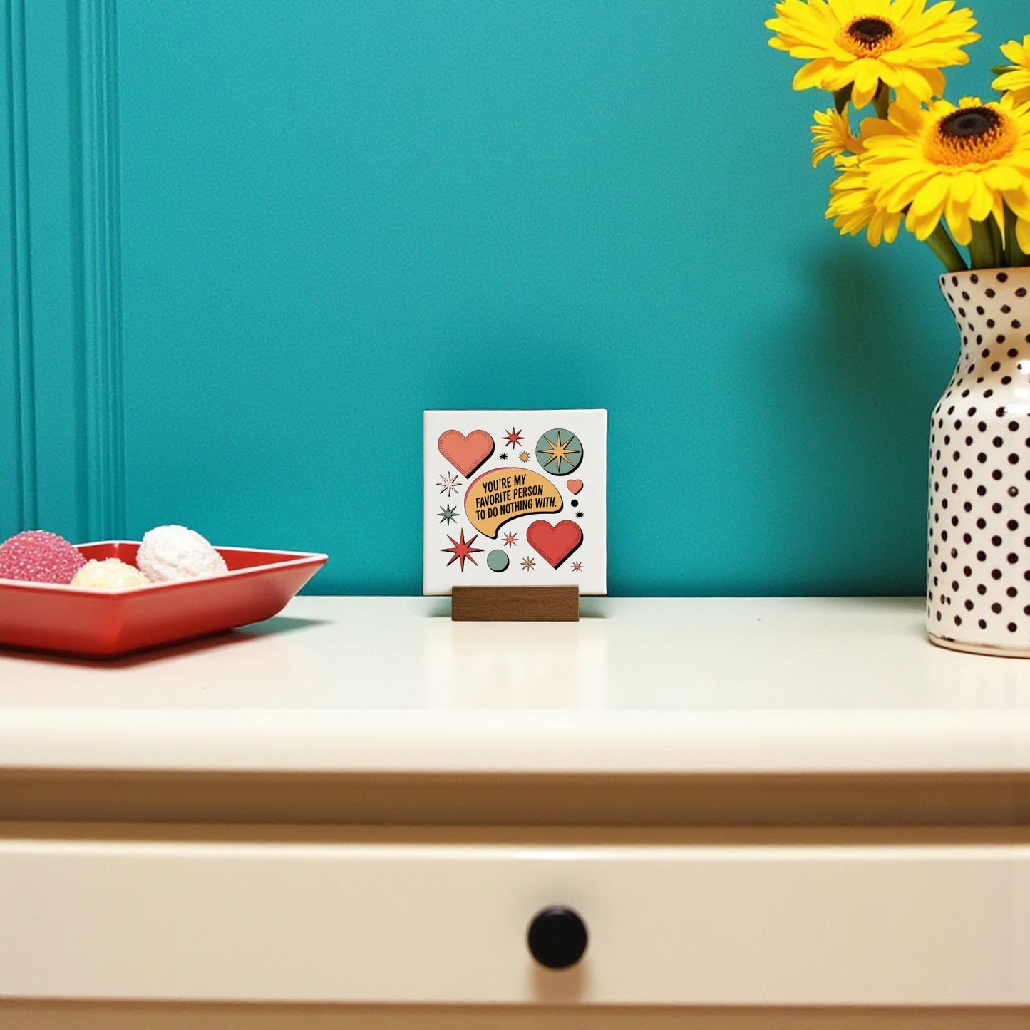 A cream-colored dresser with a black knob holds a red tray of sweets, a heart card, a white vase of yellow flowers, and the Retro Mini Love Sign by Mirabilia Boutique, against a teal wall.