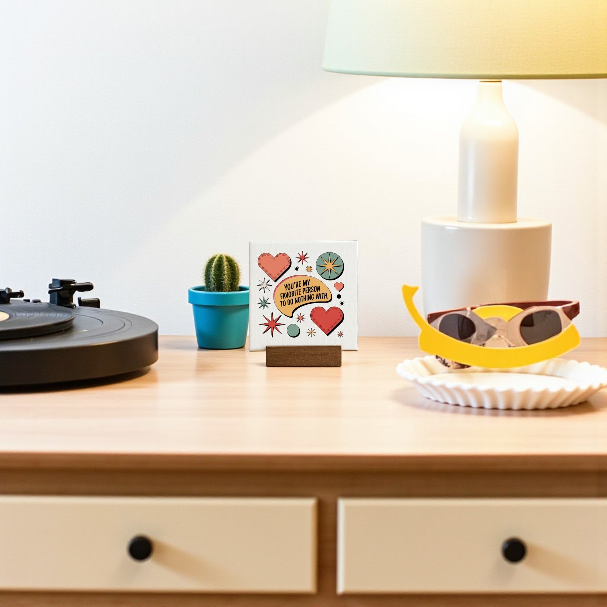 A wooden table displays Mirabilia Boutique’s Retro Mini Love Sign (“Favorite Person to Do Nothing With”) beside a record player, potted cactus, framed card with hearts, sunglasses on a tray, and a lamp—perfect for a cute or retro décor gift.