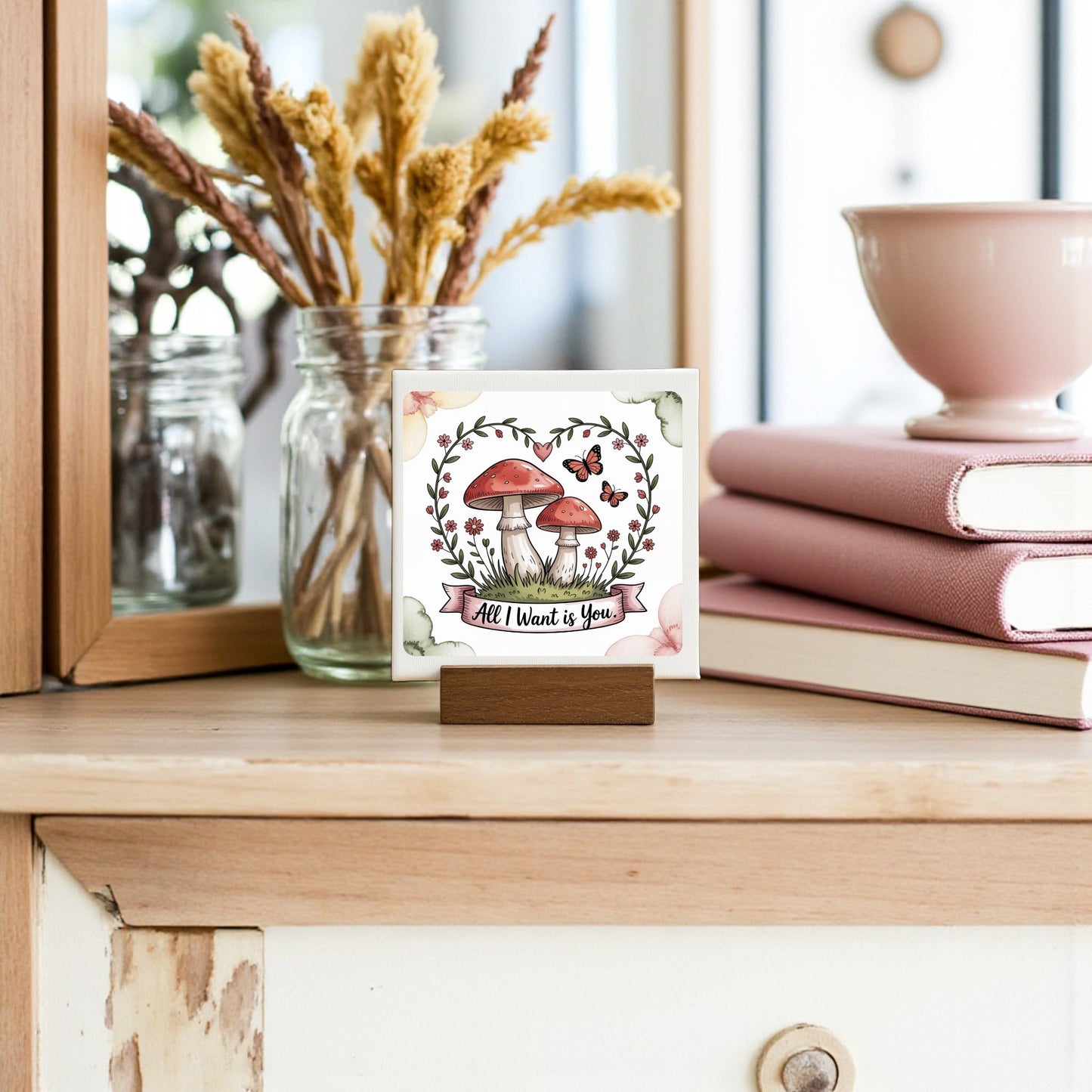 The Mini Mushroom Love Sign | All I Want Is You | 4x4 by Mirabilia Boutique, featuring illustrated mushrooms and butterflies, is a charming romantic gift or cottagecore decor for dressers, shelves, or beside pink books and dried flowers.