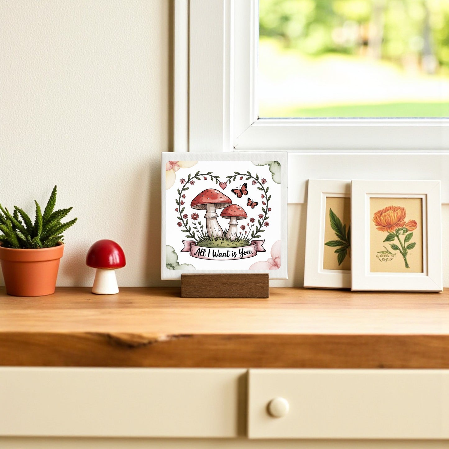 A wooden shelf features Mirabilia Boutique's Mini Mushroom Love Sign (“All I Want Is You”), a potted succulent, framed mushroom and flower prints—perfect cottagecore decor—set against a window with lush greenery outside.