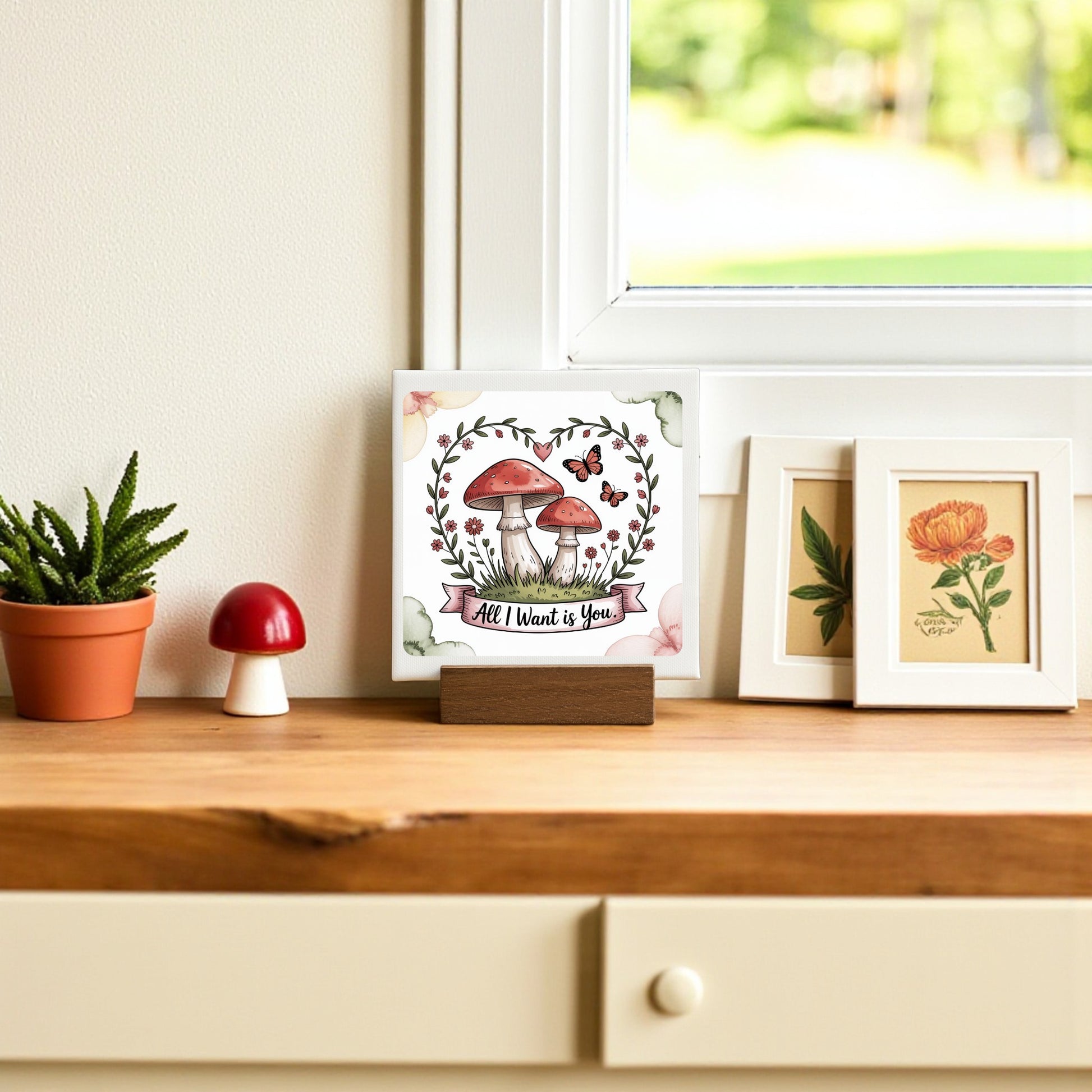 A wooden shelf features Mirabilia Boutique's Mini Mushroom Love Sign (“All I Want Is You”), a potted succulent, framed mushroom and flower prints—perfect cottagecore decor—set against a window with lush greenery outside.