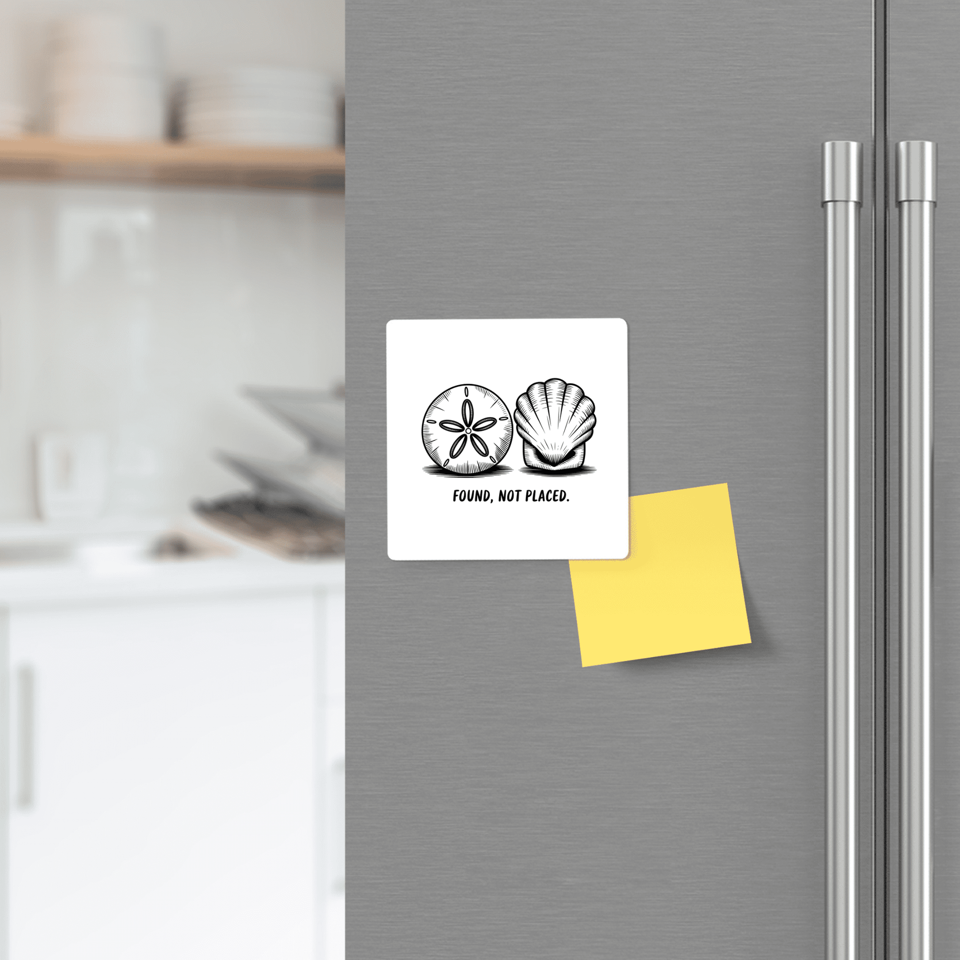 A Mirabilia Boutique "Found, Not Placed" Magnet – Minimalist Coastal Quote with a hand-drawn shell decorates the fridge above a yellow sticky note, set against kitchen shelves and subtle beach-themed accents.