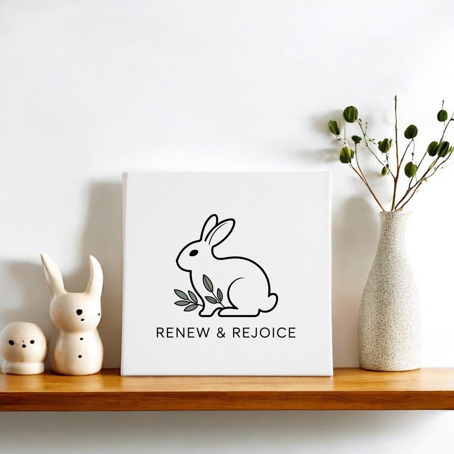 A wooden shelf holds the Mirabilia Boutique Renew & Rejoice Bunny Canvas 8x8 Spring Wall Art, ceramic bunny figurines, and a tall vase of green branches—minimalist Easter decor against a white wall.
