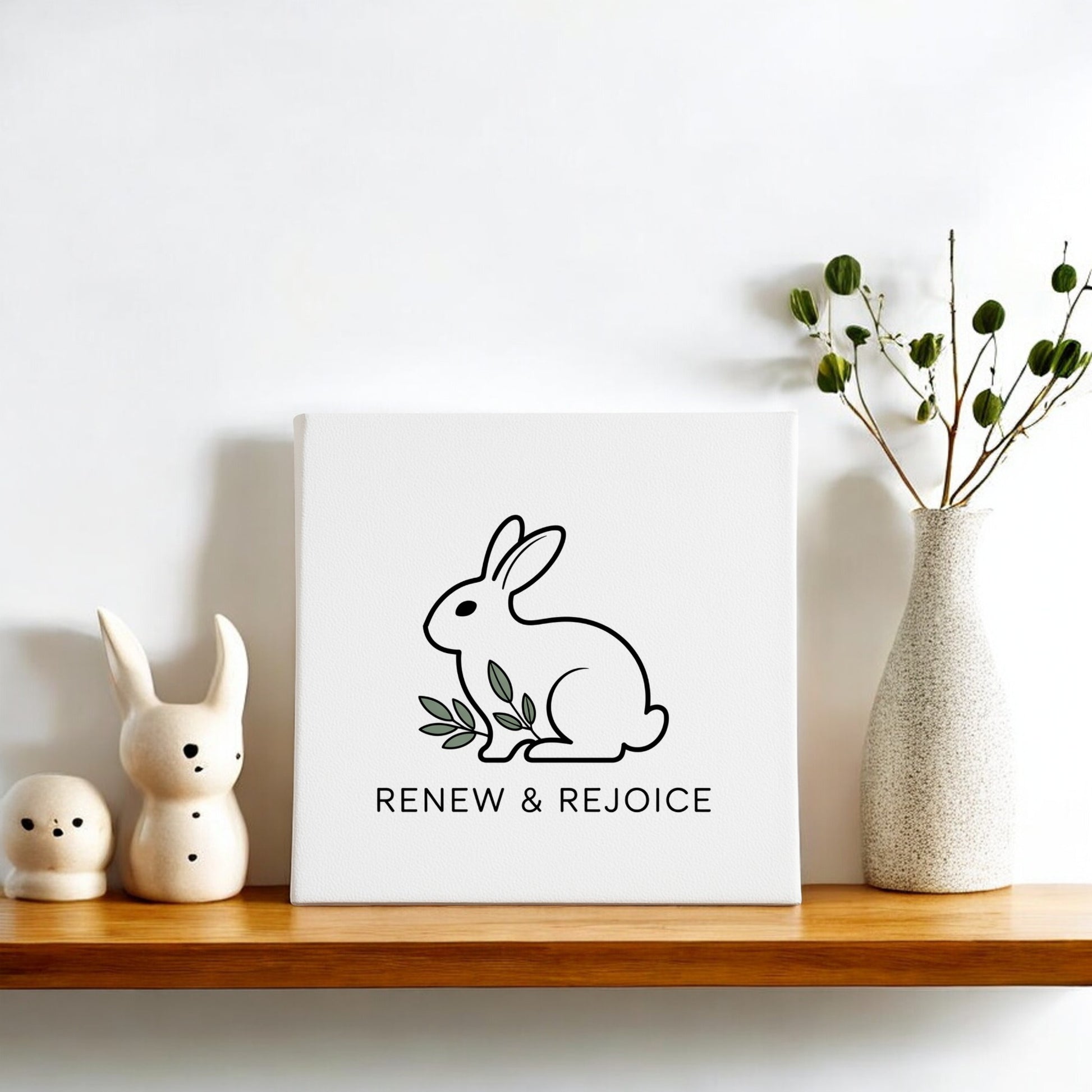 A wooden shelf holds the Mirabilia Boutique Renew & Rejoice Bunny Canvas 8x8 Spring Wall Art, ceramic bunny figurines, and a tall vase of green branches—minimalist Easter decor against a white wall.