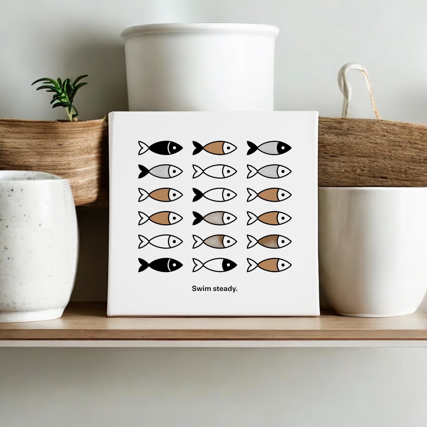 The Mirabilia Boutique Swim Steady Canvas – Minimalist Neutral Wall Art, 8x8, features simple black, white, and brown fish illustrations. Displayed on a shelf between two cups with a plant behind, it offers gentle encouragement in a modern style.