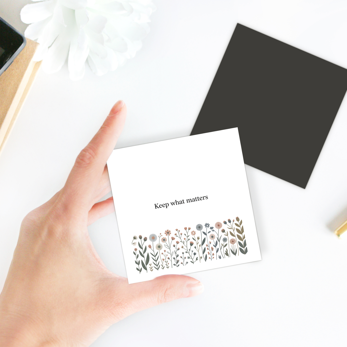 A hand holds the Mirabilia Boutique "Keep What Matters" Floral Magnet. Surrounding it are a black square paper, white flower, and office supplies, creating a charming cottage décor vibe on the white surface.