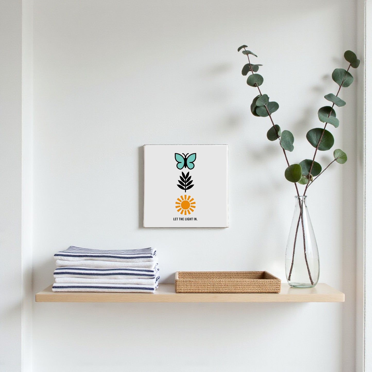 A minimalist shelf displays folded striped towels, a wicker tray, and a glass vase with eucalyptus. Above hangs the Let the Light In Canvas – Butterfly Sun Wall Art by Mirabilia Boutique, adding inspiring decor.