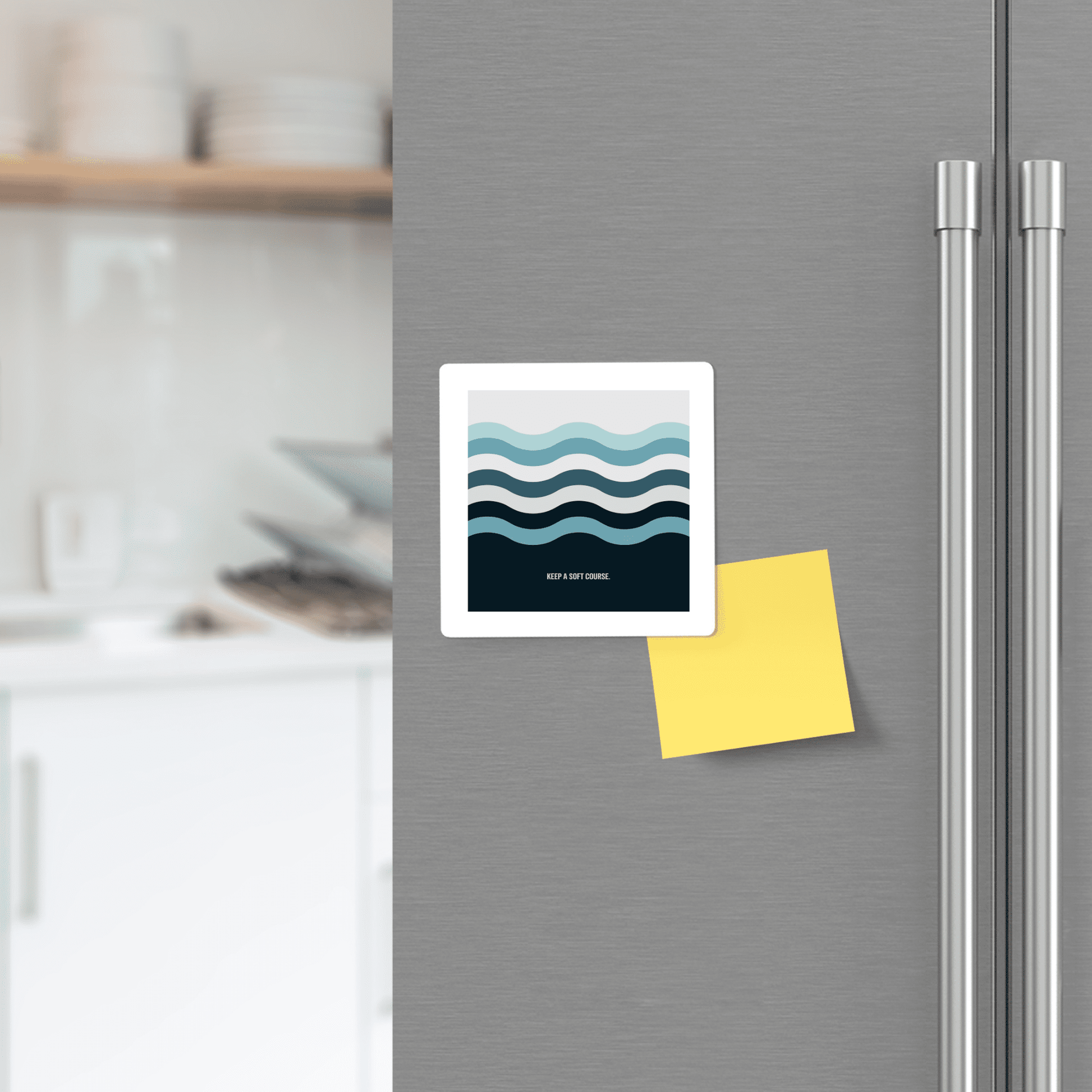 A stainless steel fridge door displays the Mirabilia Boutique Keep a Soft Course Magnet—a minimalist, ocean-inspired square with wavy blue lines—beside a yellow sticky note. Blurred kitchen counter and dishes appear in the background.